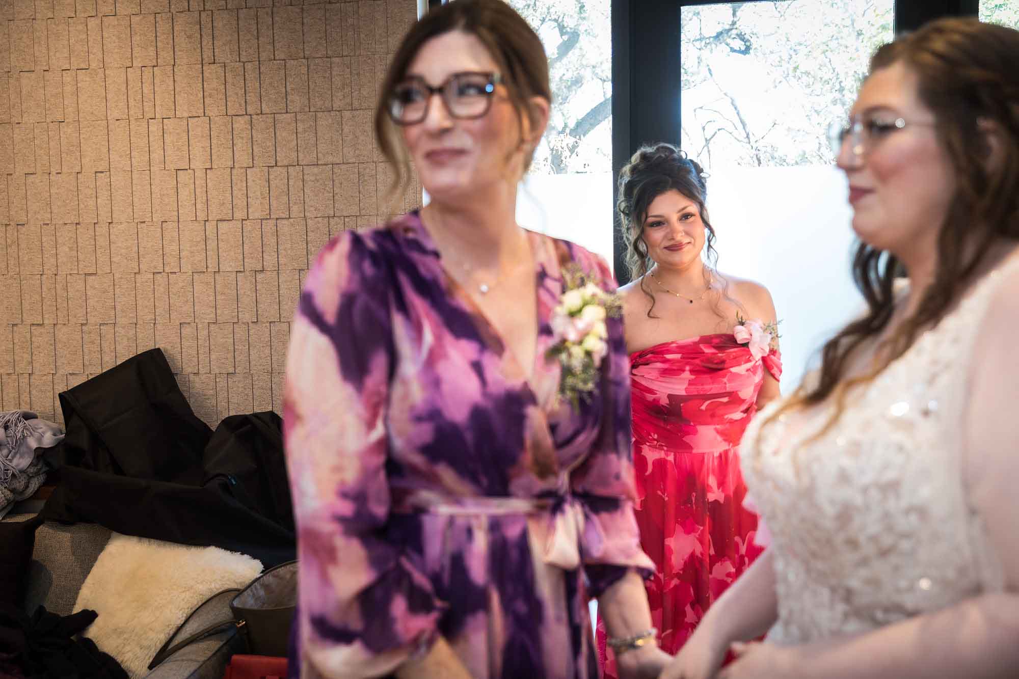 Bride and mother wearing purple floral dress standing in front of woman wearing red floral dress and window before a San Antonio Botanical Garden wedding ceremony
