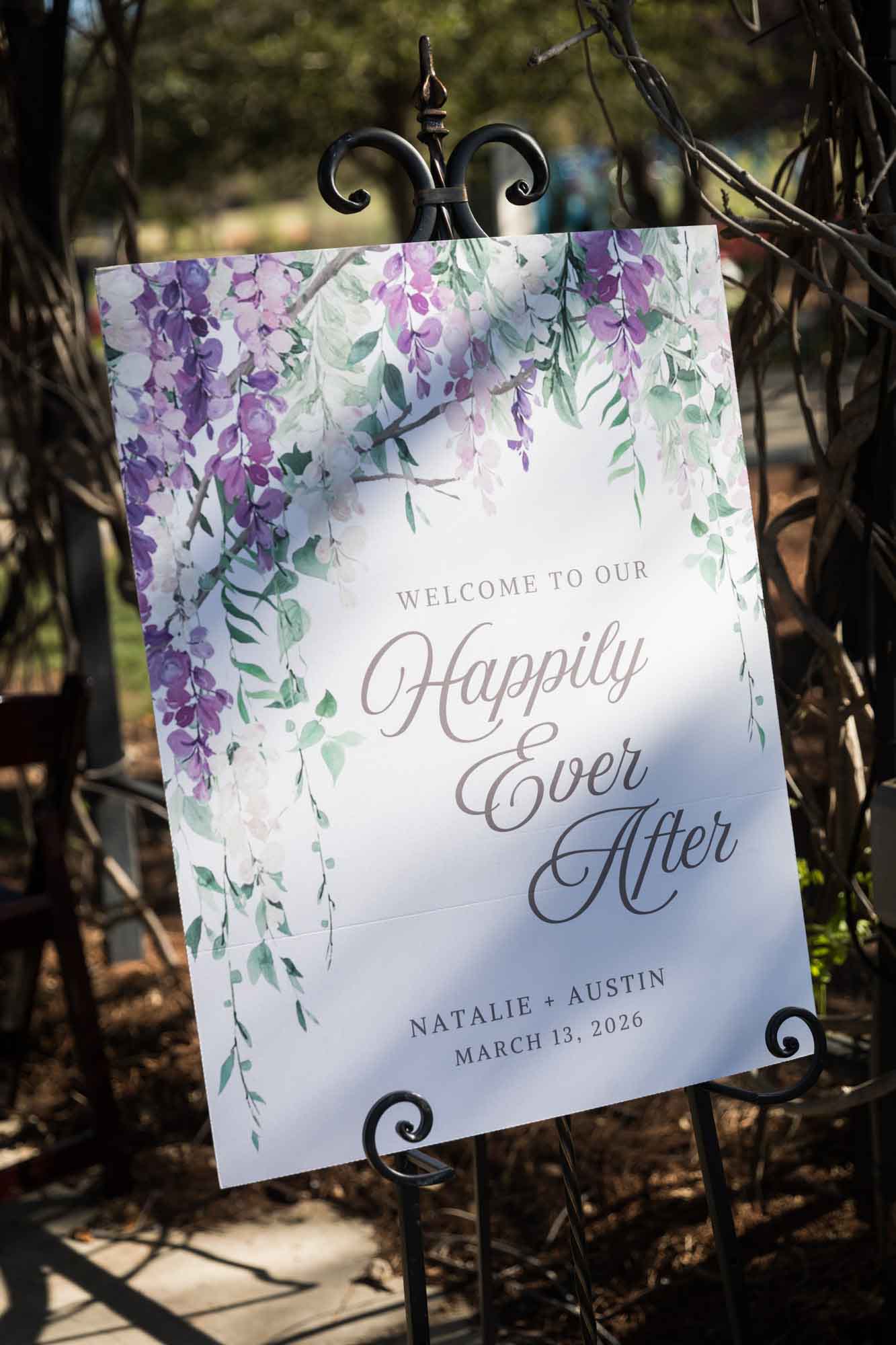 White poster with purple wisteria painted in corner and sign saying 'Welcome to Our Happily Ever After' during a San Antonio Botanical Garden wedding ceremony