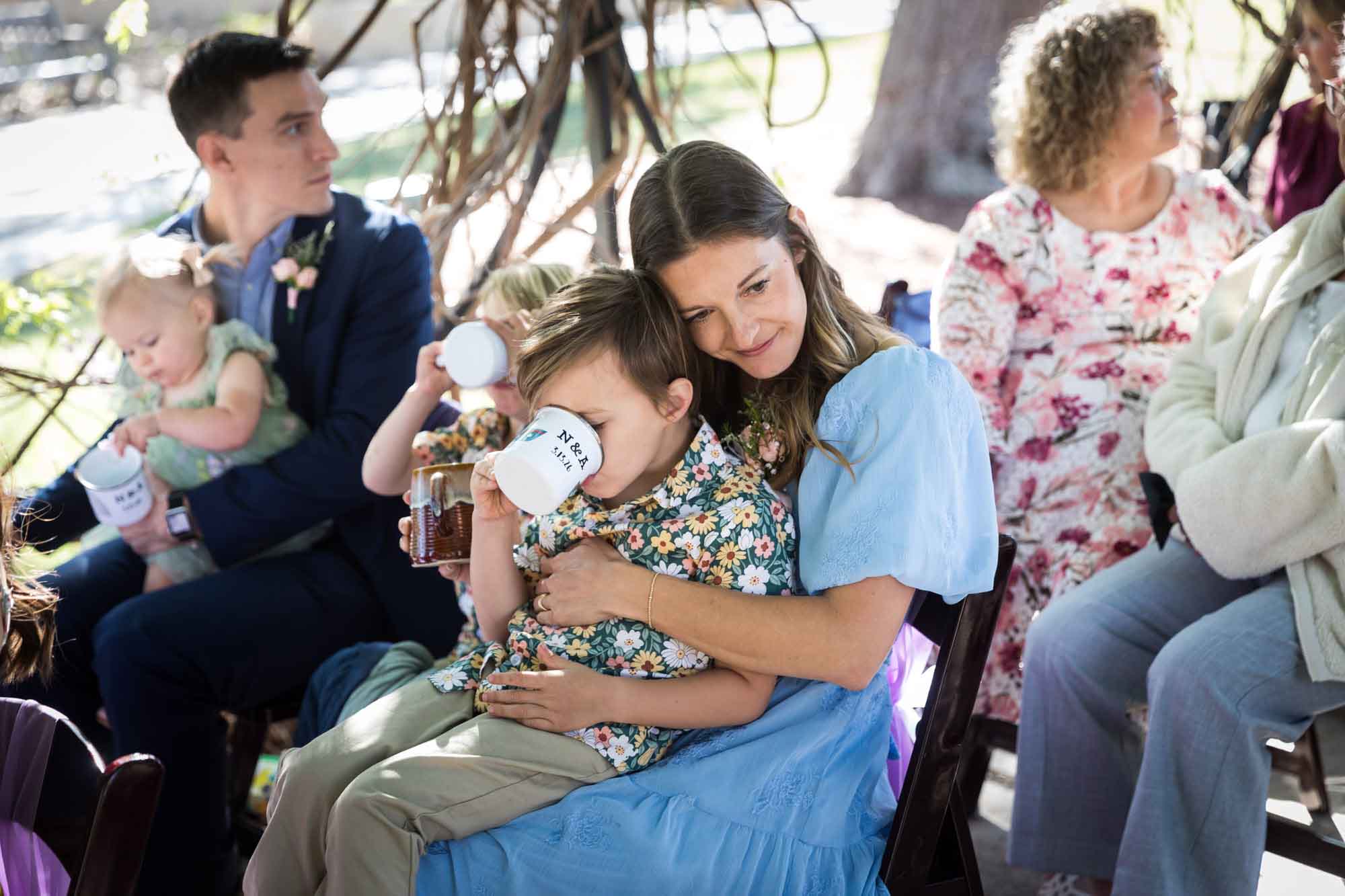 Little boy drinking from a mug while sitting in his mother's lap sitting in chairs during a San Antonio Botanical Garden wedding ceremony