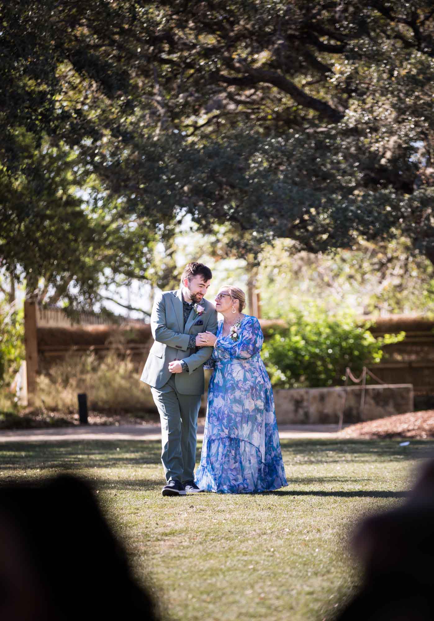 Mother and son holding arms and walking across lawn during a San Antonio Botanical Garden wedding ceremony