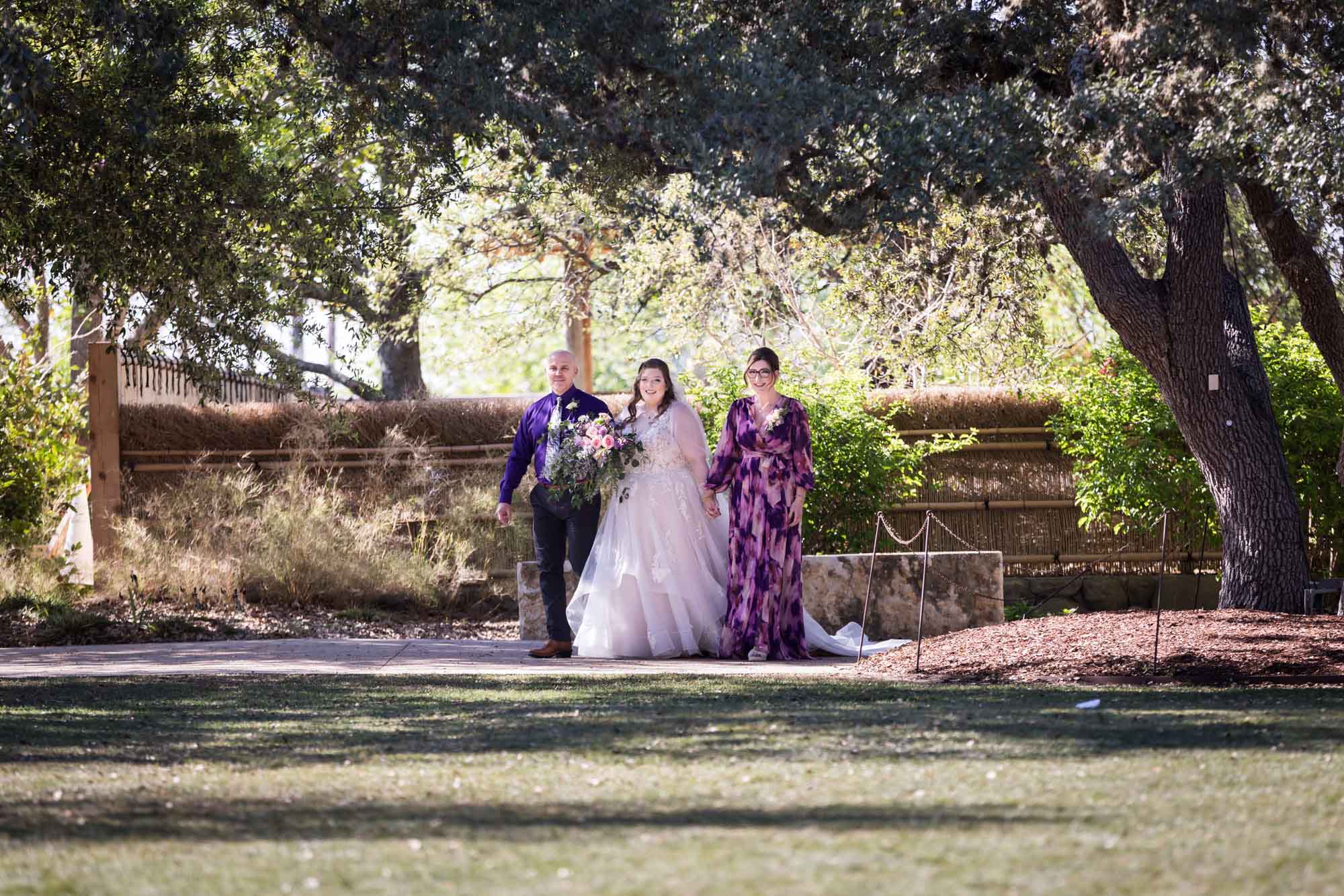 Bride holding hands with mother and father walking across lawn during a San Antonio Botanical Garden wedding ceremony