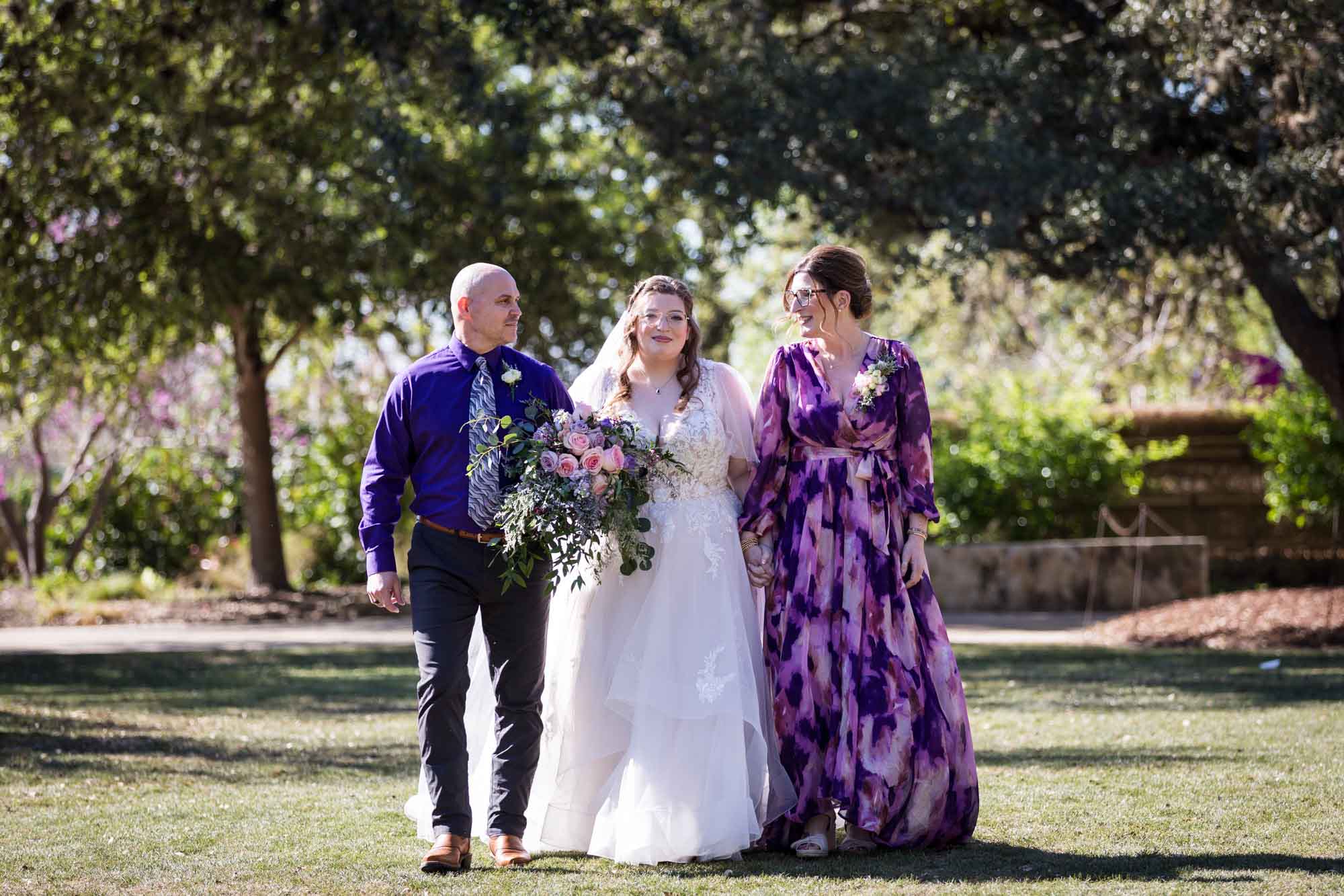 Bride walking with parents across lawn during a San Antonio Botanical Garden wedding ceremony