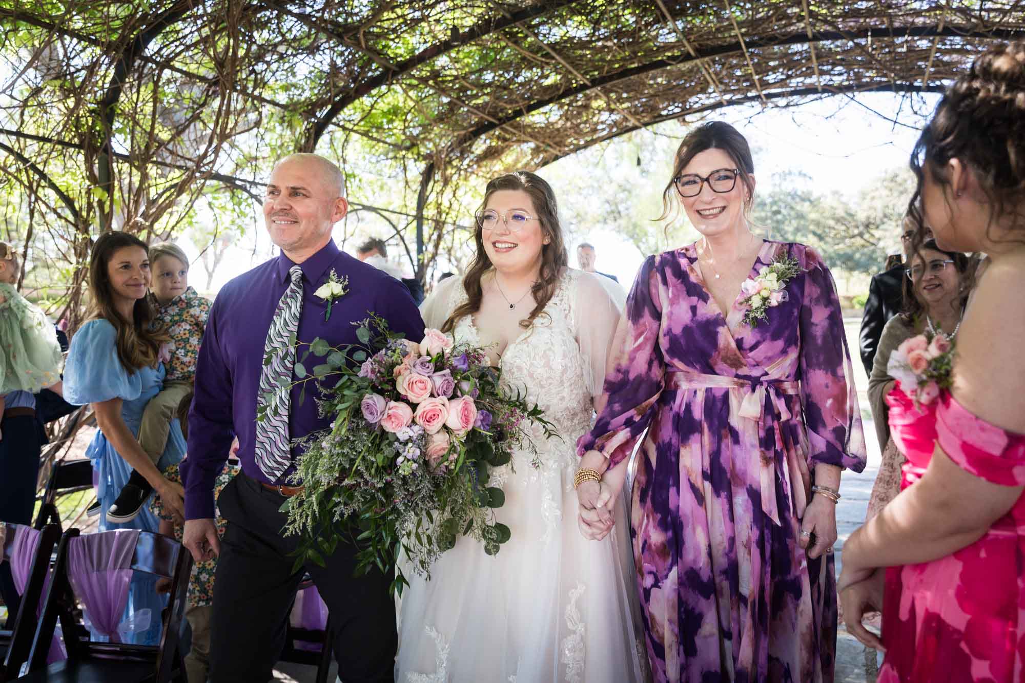 Bride walking with mother and father in front of other guests under Wisteria Arbor during a San Antonio Botanical Garden wedding ceremony