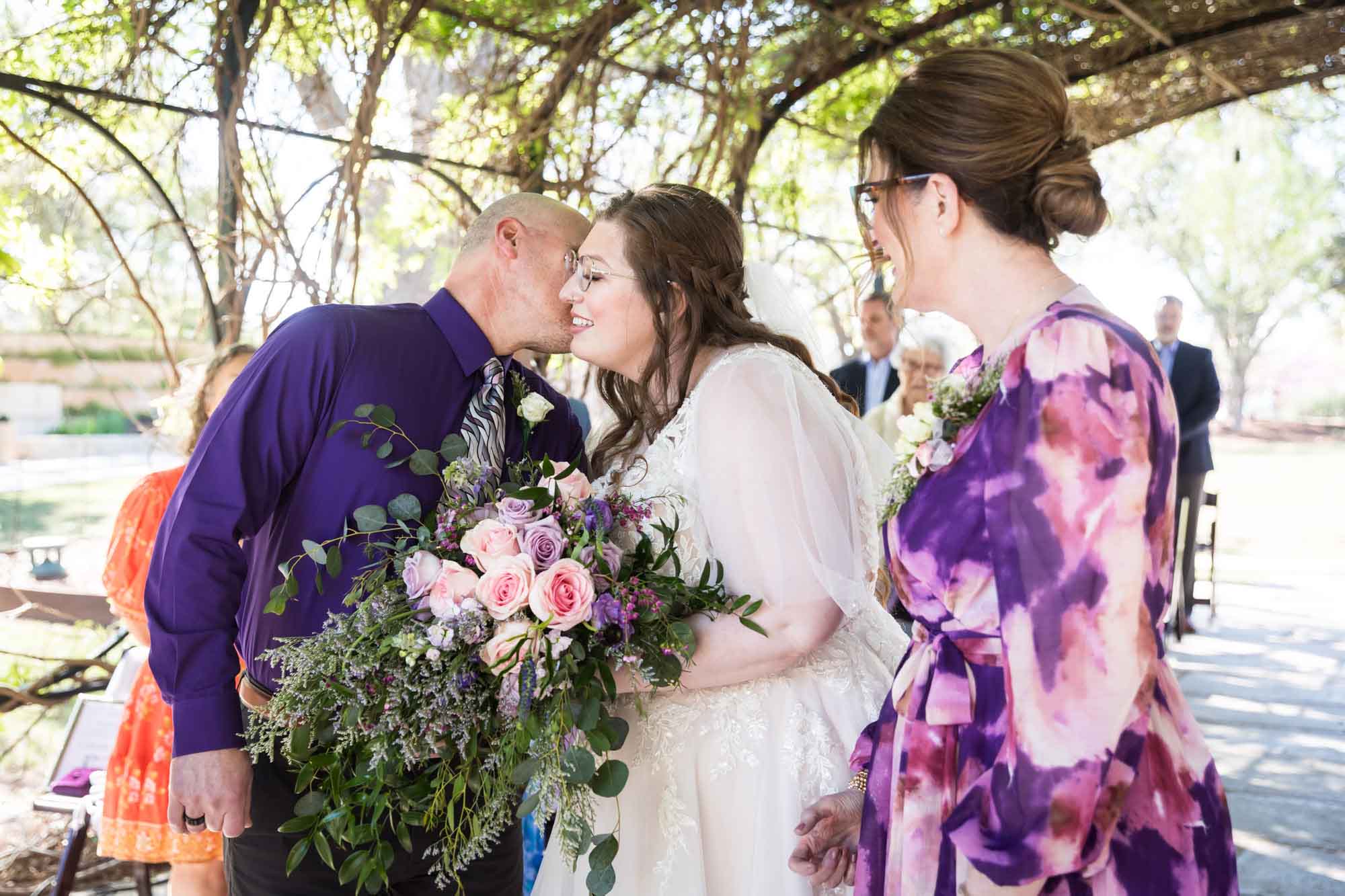 Father kissing bride on the cheek beside mother under Wisteria Arbor during a San Antonio Botanical Garden wedding ceremony