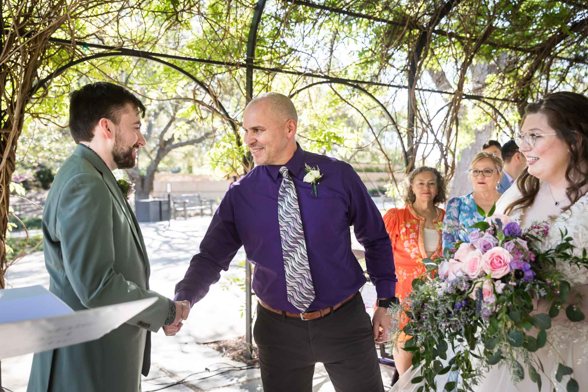 Father shaking hands with groom in front of bride holding bouquet in front of guests during a San Antonio Botanical Garden wedding ceremony