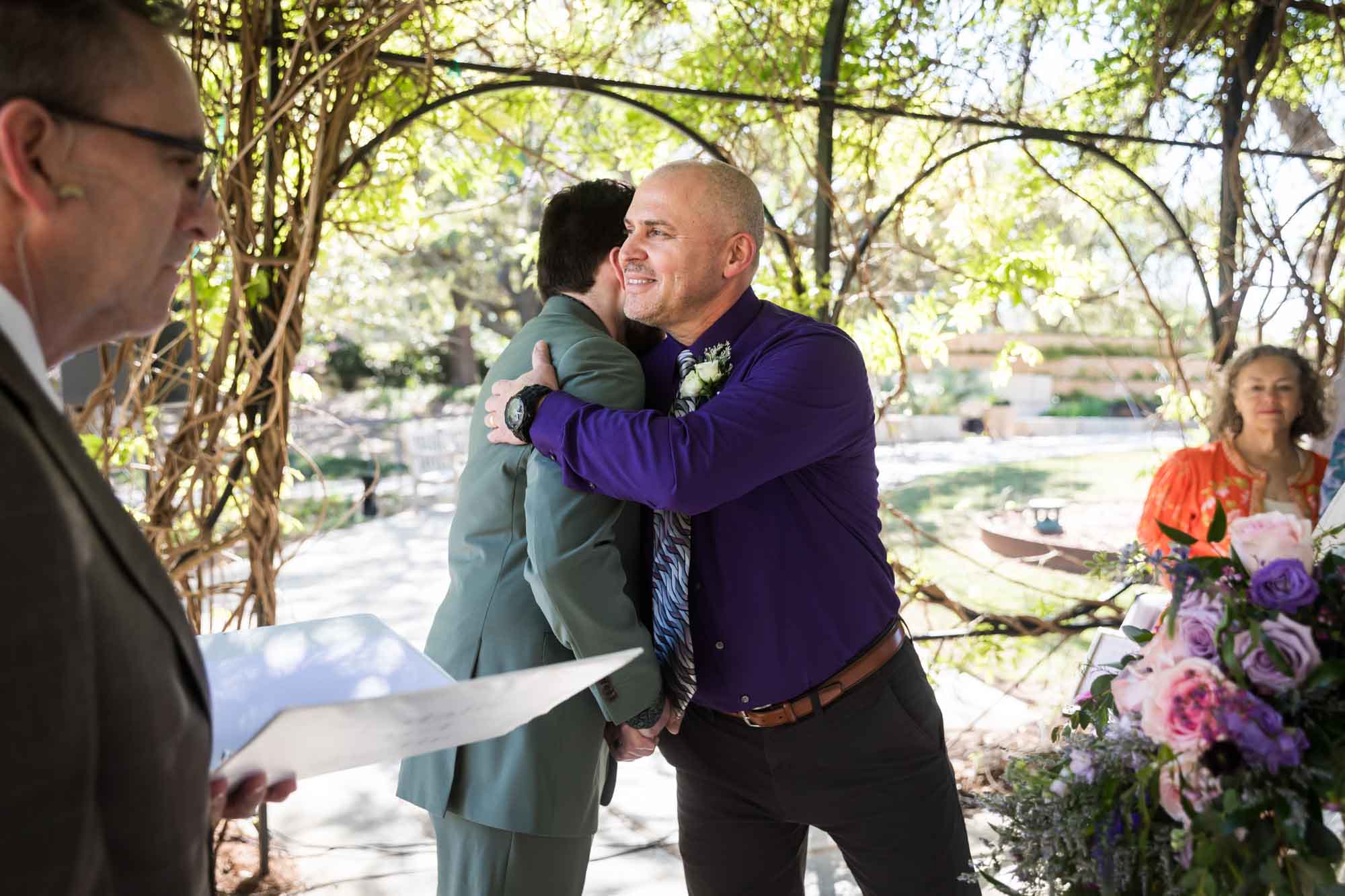 Father hugging groom in front of officiant and guests during a San Antonio Botanical Garden wedding ceremony