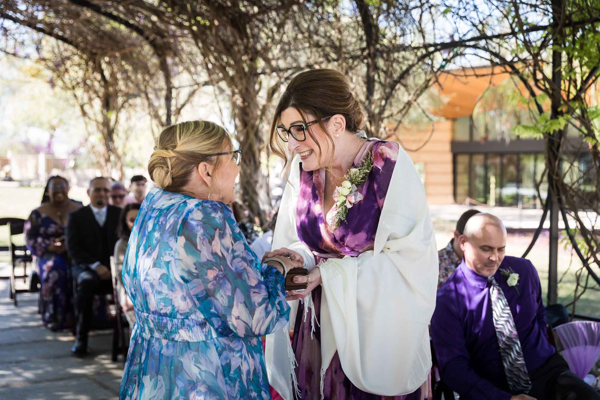 Two mothers holding wooden box in front of guests under Wisteria Arbor during a San Antonio Botanical Garden wedding ceremony