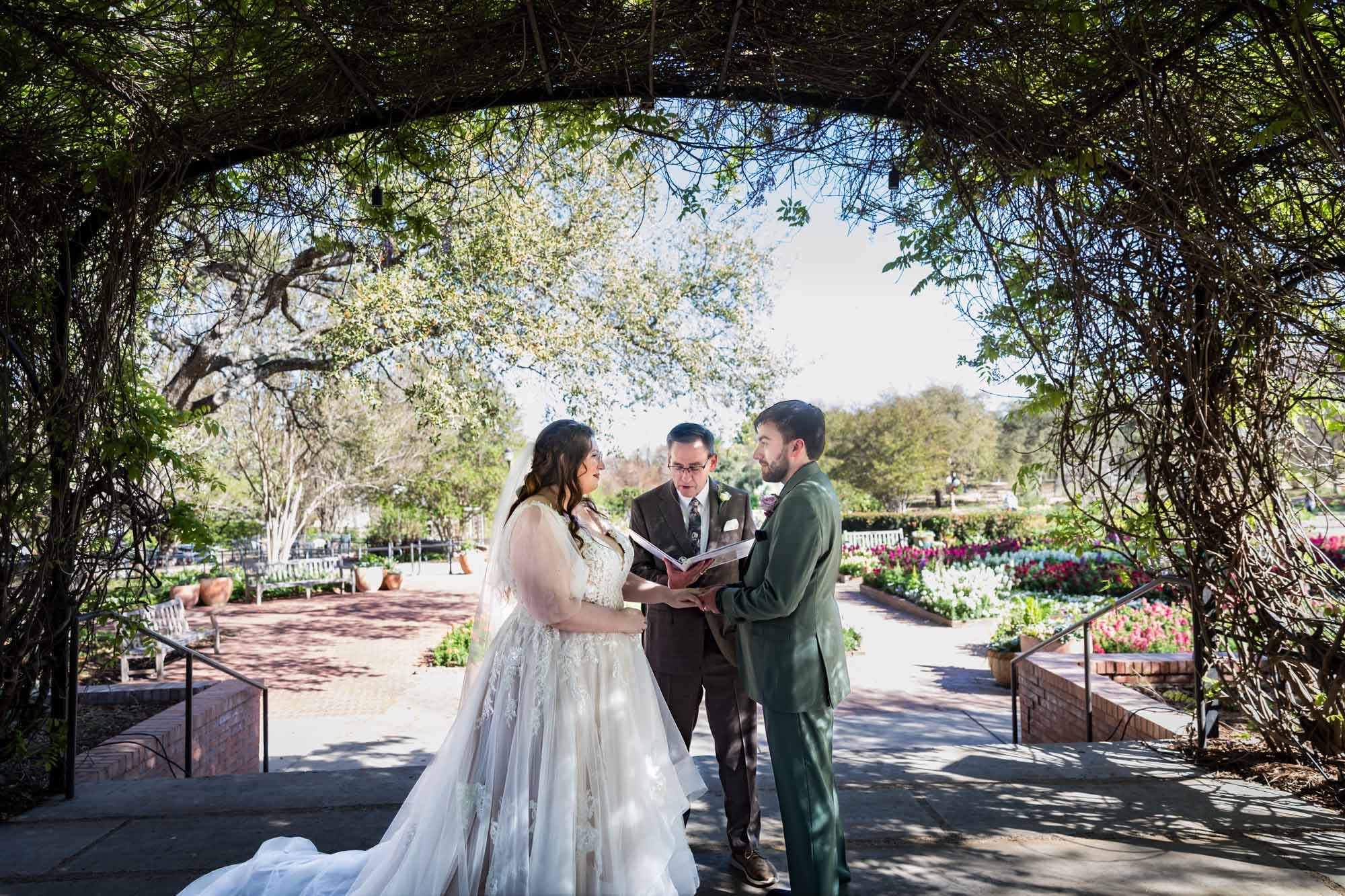 Bride and groom holding hands in front of officiant under Wisteria Arbor during a San Antonio Botanical Garden wedding ceremony