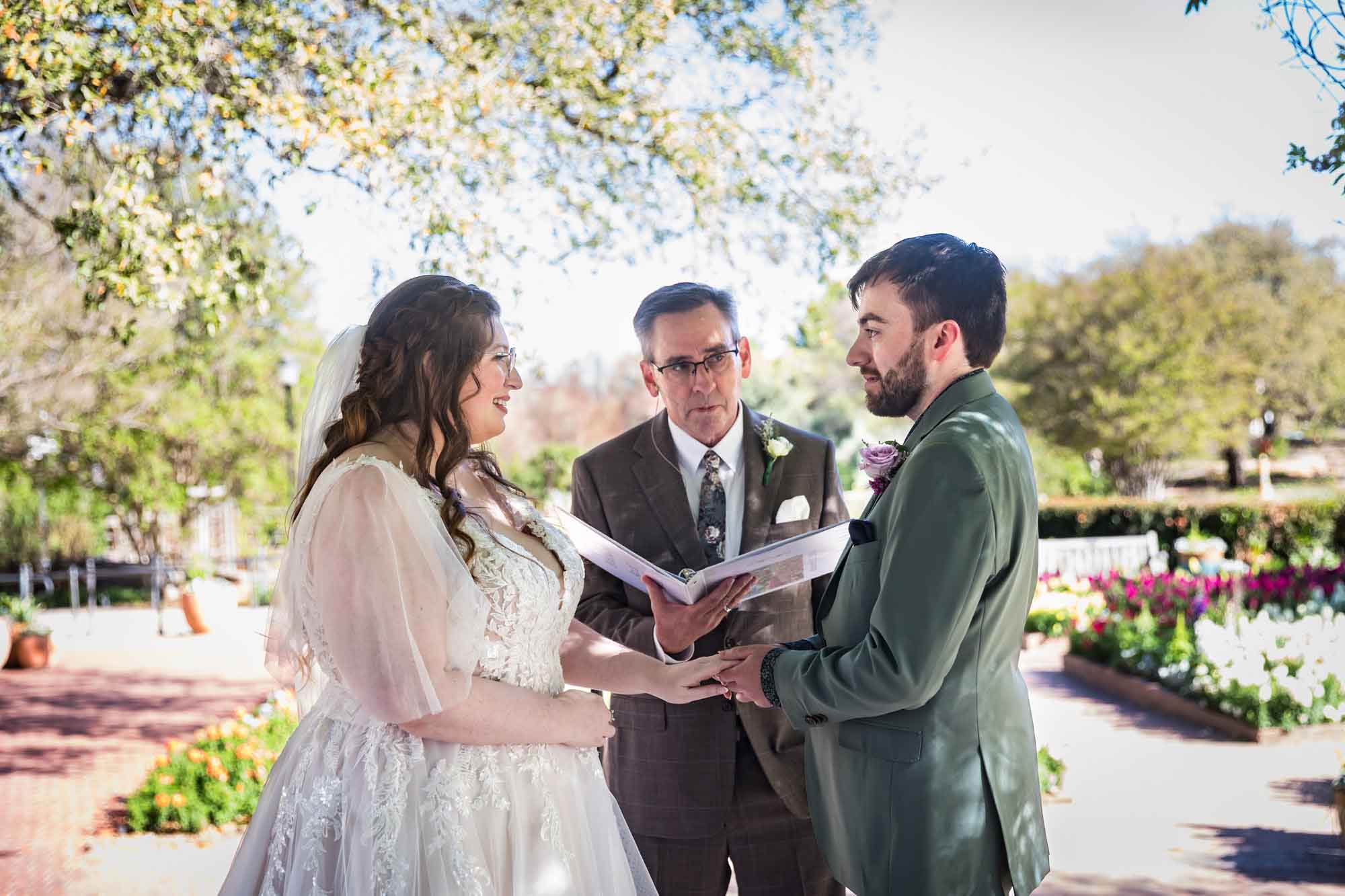 Bride and groom holding hands in front of officiant holding notebook during a San Antonio Botanical Garden wedding ceremony
