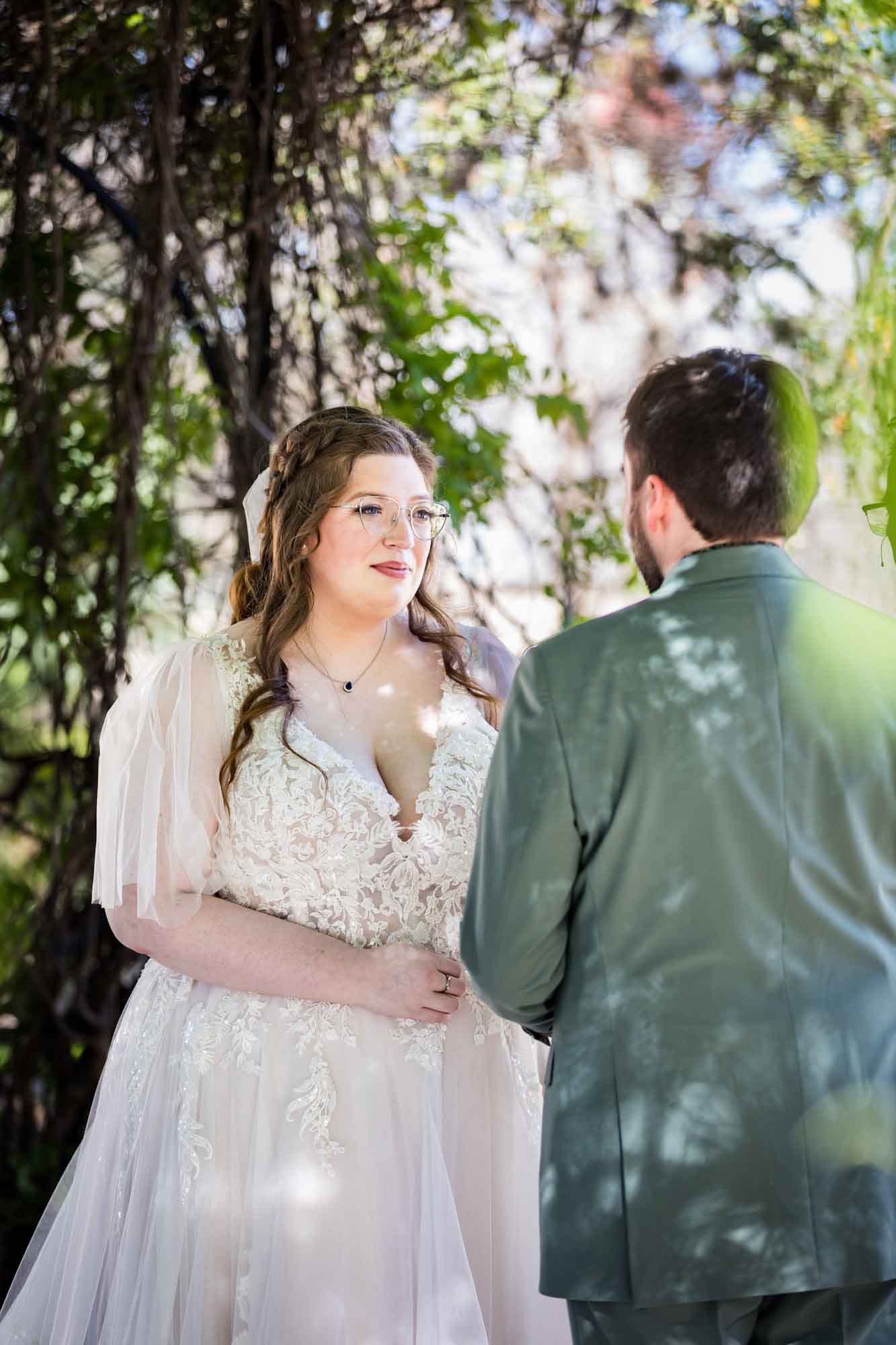 Bride listening to groom wearing green suit under Wisteria Arbor during a San Antonio Botanical Garden wedding ceremony