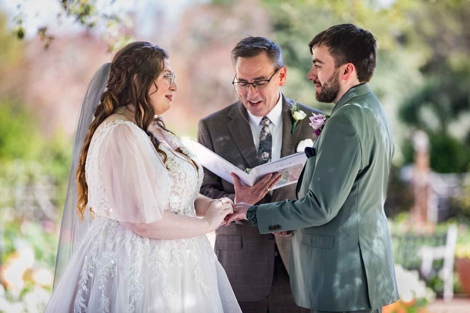 Bride putting ring on groom's finger in front of officiant holding notebook during a San Antonio Botanical Garden wedding ceremony during a San Antonio Botanical Garden wedding ceremony