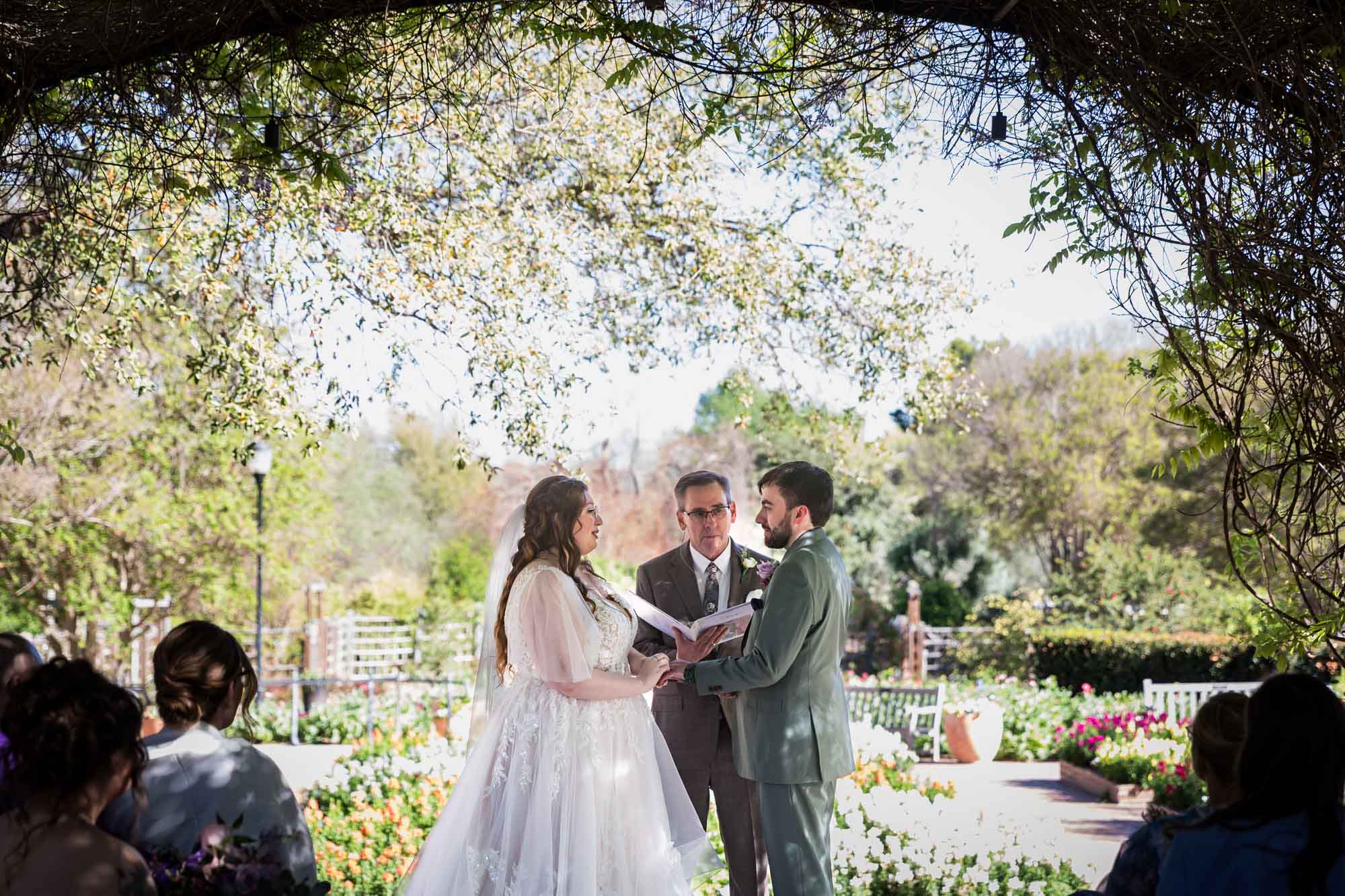 Bride and groom holding hands in front of officiant holding notebook in front of flower gardens during a San Antonio Botanical Garden wedding ceremony