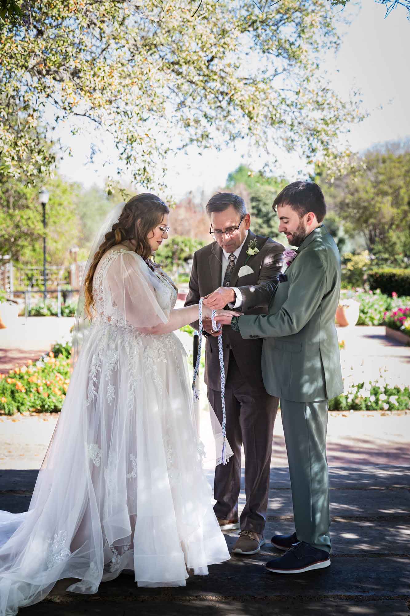 Officiant wrapping bride and groom with braided rope during handfasting ritual in front of flower gardens during a San Antonio Botanical Garden wedding ceremony