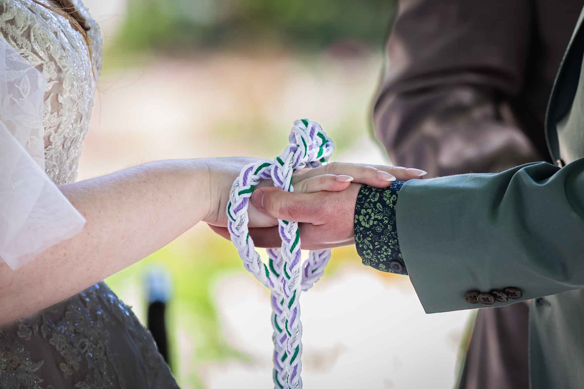 Close up of man and woman holding hands while wrapped in white, purple, and green braided rope during handfasting ceremony