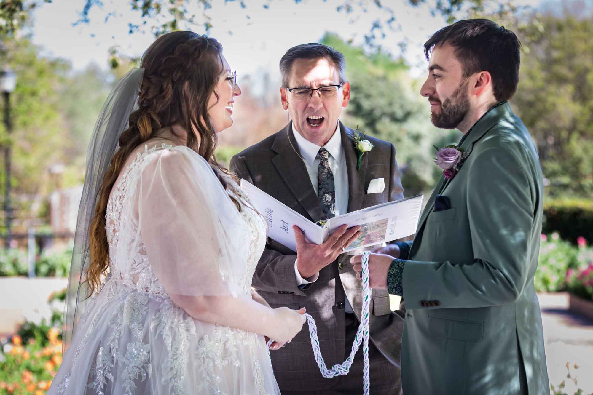 Bride and groom holding braided rope in front of officiant standing with open mouth during handfasting ceremony during a San Antonio Botanical Garden wedding ceremony