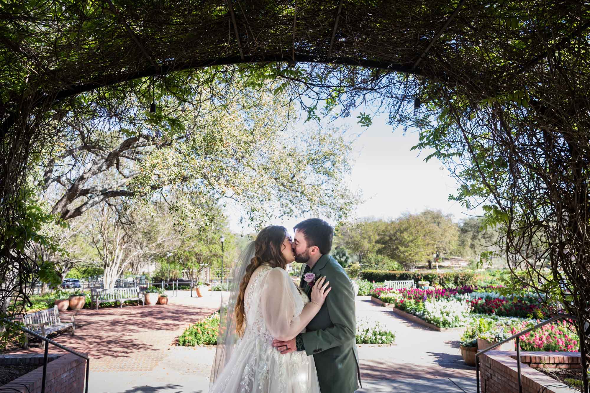 Bride and groom kissing under Wisteria Arbor in front of colorful gardens during a San Antonio Botanical Garden wedding ceremony
