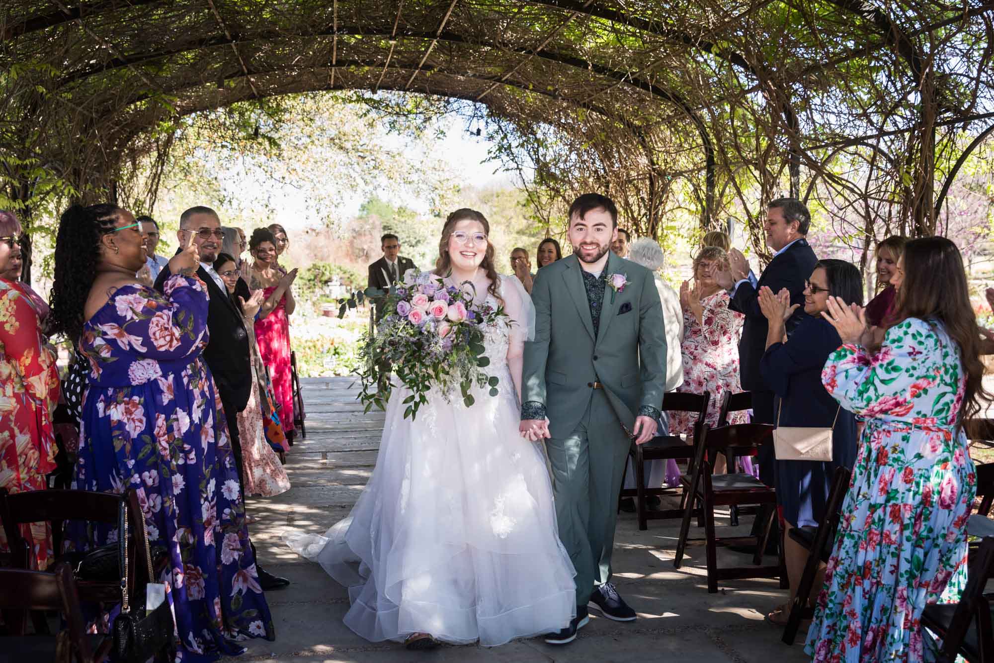 Bride holding colorful bouquet walking with groom down aisle in front of guests under Wisteria Arbor during a San Antonio Botanical Garden wedding ceremony