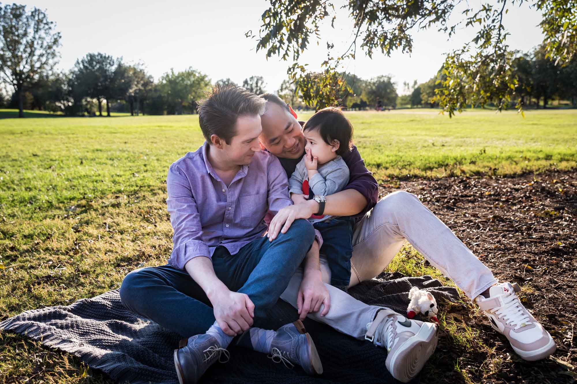 Two fathers playing with little boy while sitting on grass during a Mueller Lake Park family portrait