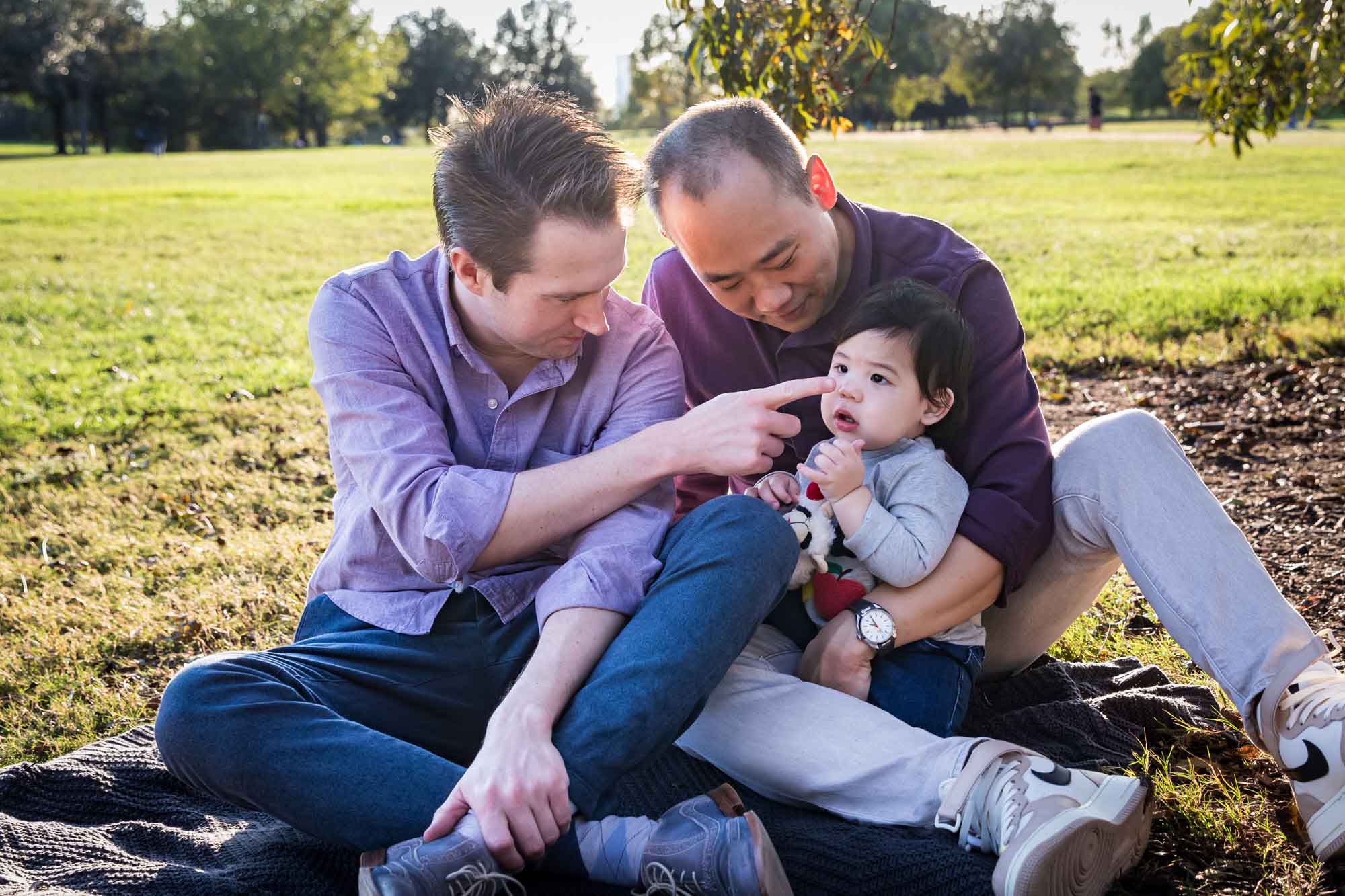 Two fathers playing with little boy while sitting on grass during a Mueller Lake Park family portrait