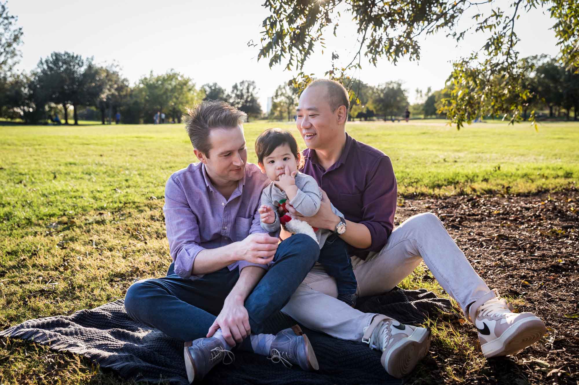 Two fathers playing with little boy while sitting on grass during a Mueller Lake Park family portrait