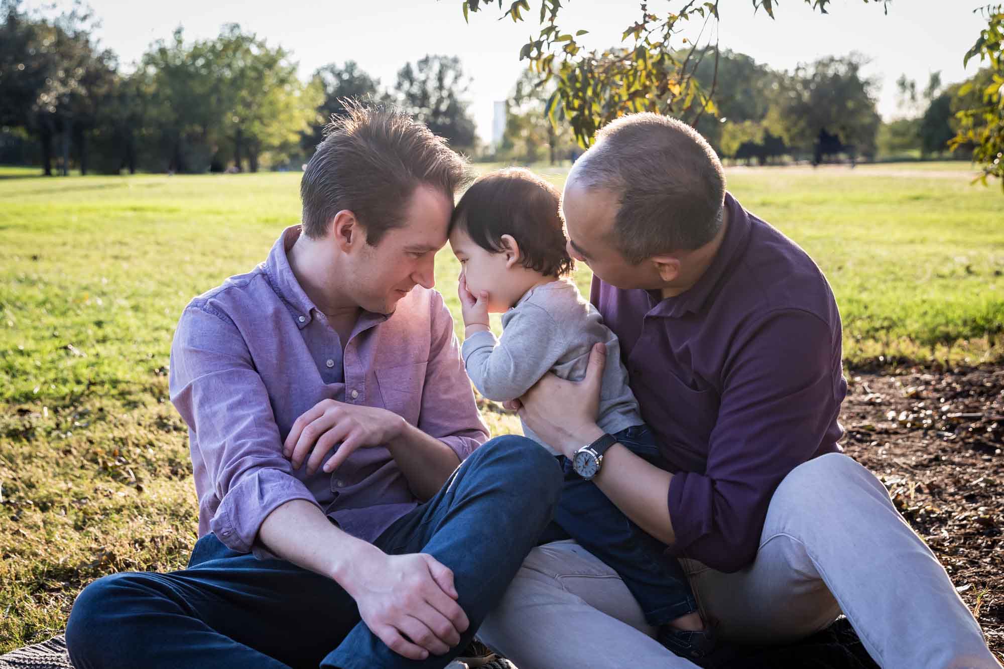 Two fathers playing with little boy while sitting on grass during a Mueller Lake Park family portrait