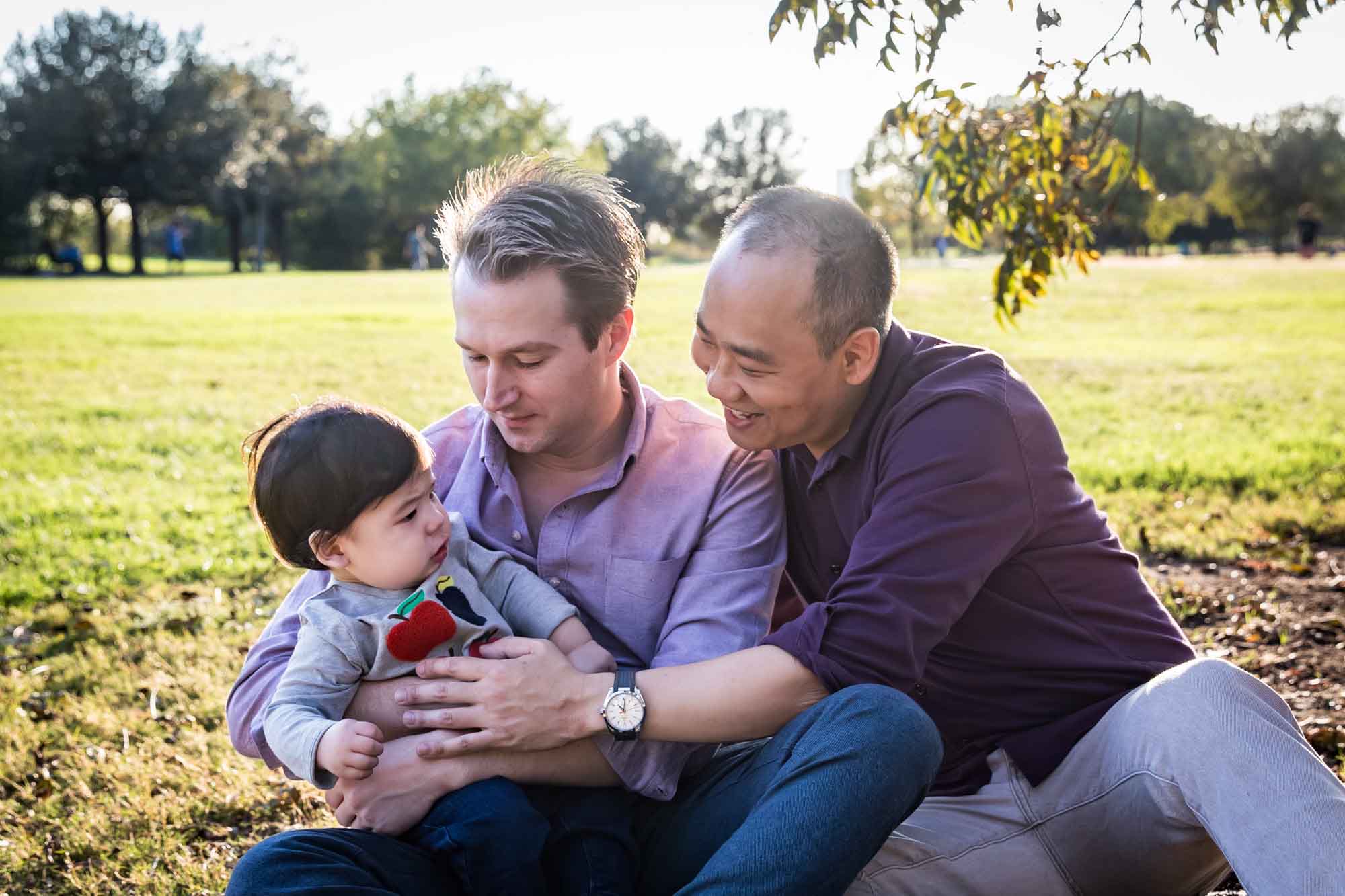 Two fathers playing with little boy while sitting on grass during a Mueller Lake Park family portrait