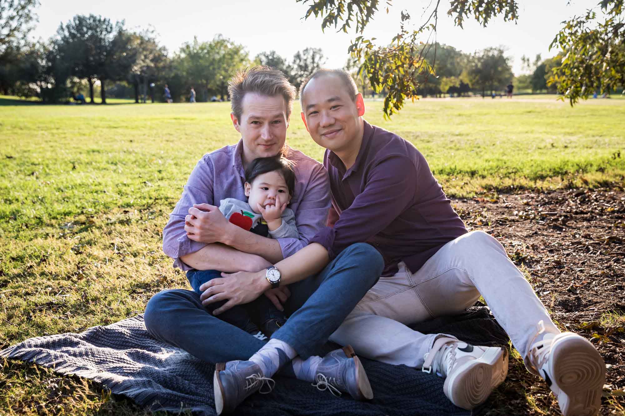Two fathers playing with little boy while sitting on grass during a Mueller Lake Park family portrait