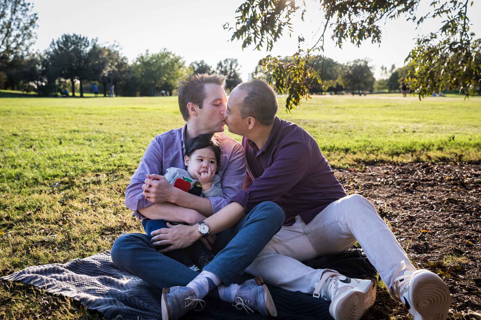 Two fathers playing with little boy while sitting on grass during a Mueller Lake Park family portrait