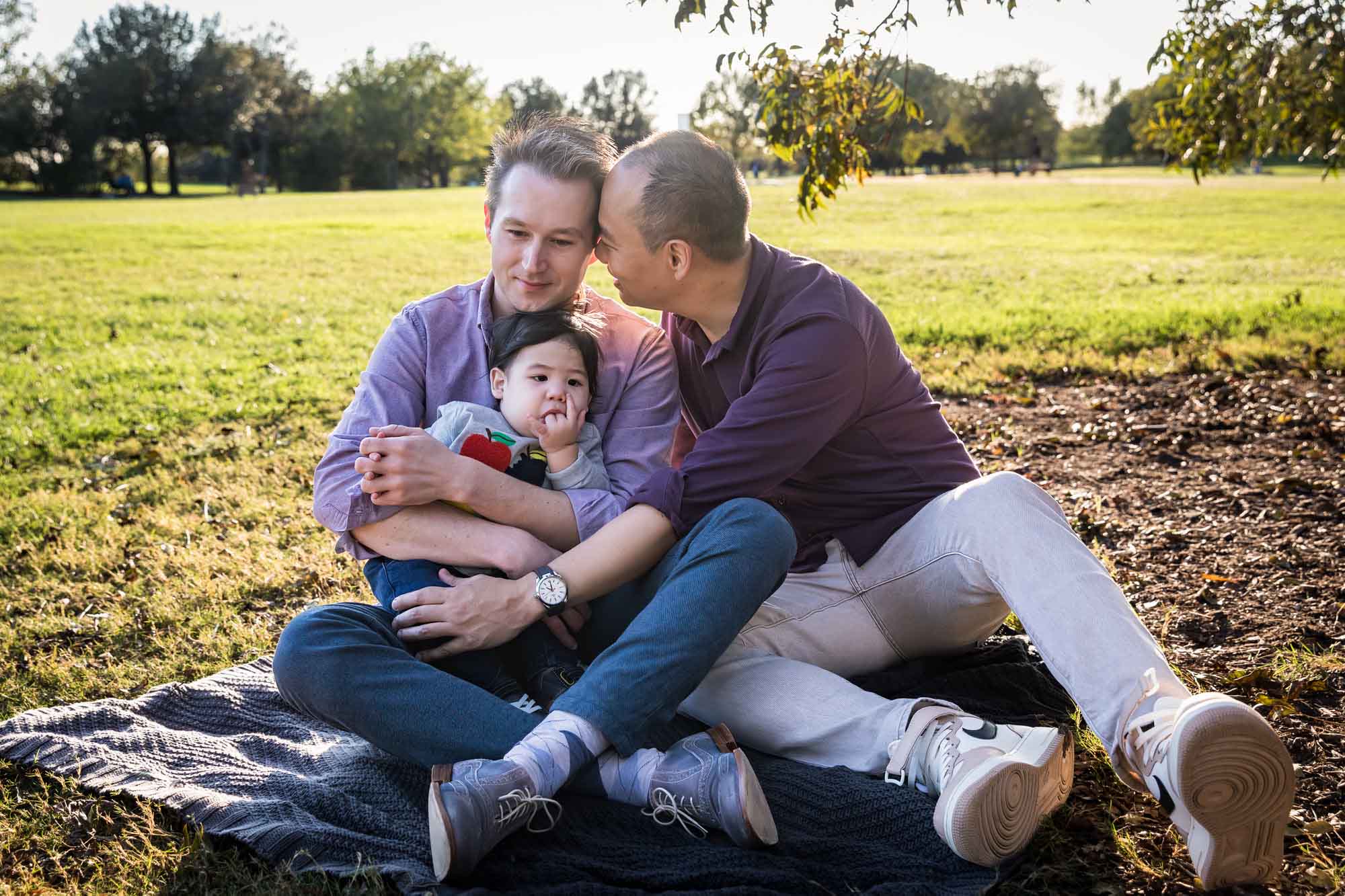 Two fathers playing with little boy while sitting on grass during a Mueller Lake Park family portrait