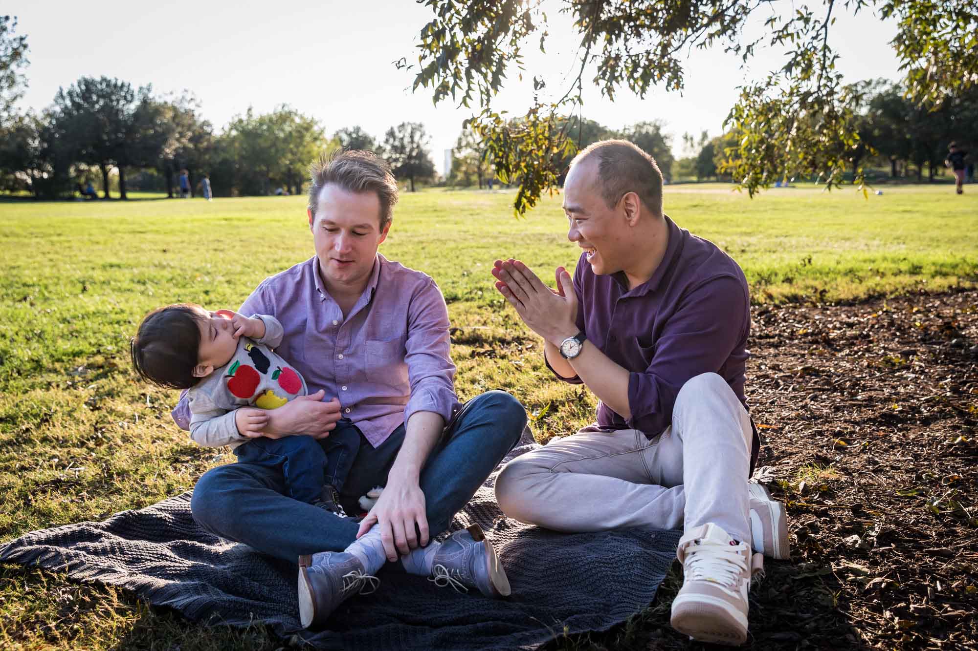 Two fathers playing with little boy while sitting on grass during a Mueller Lake Park family portrait