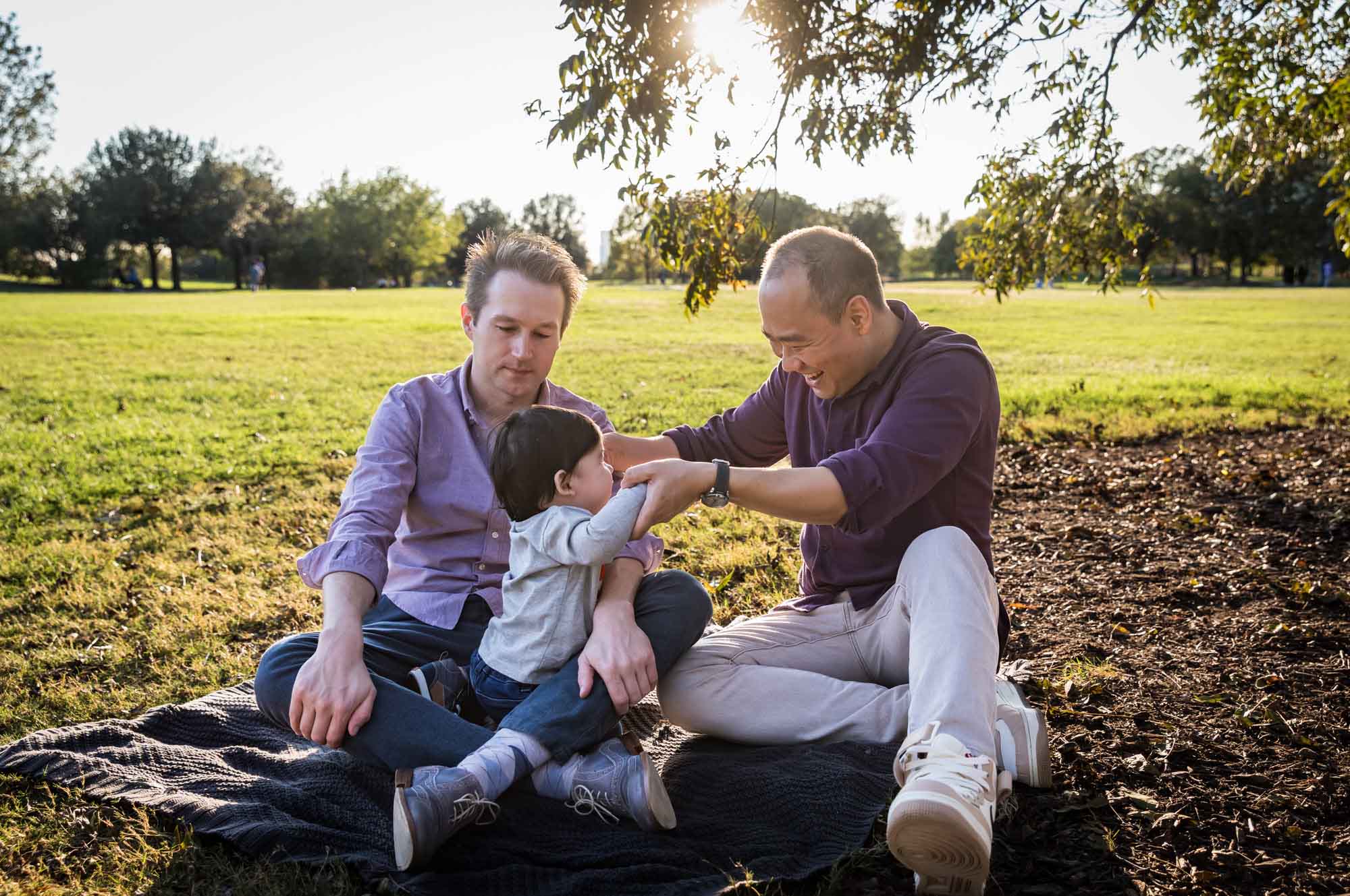 Two fathers playing with little boy while sitting on grass during a Mueller Lake Park family portrait
