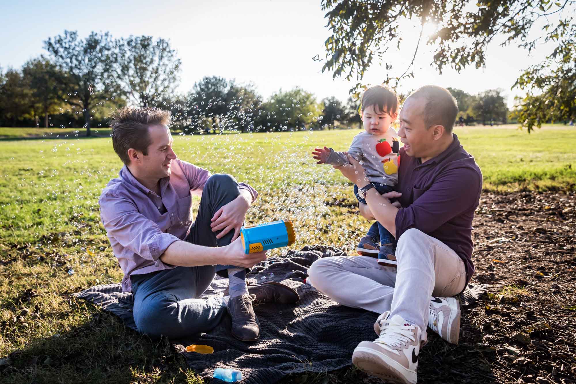 Two fathers playing with little boy surrounded by bubbles while sitting on grass during a Mueller Lake Park family portrait