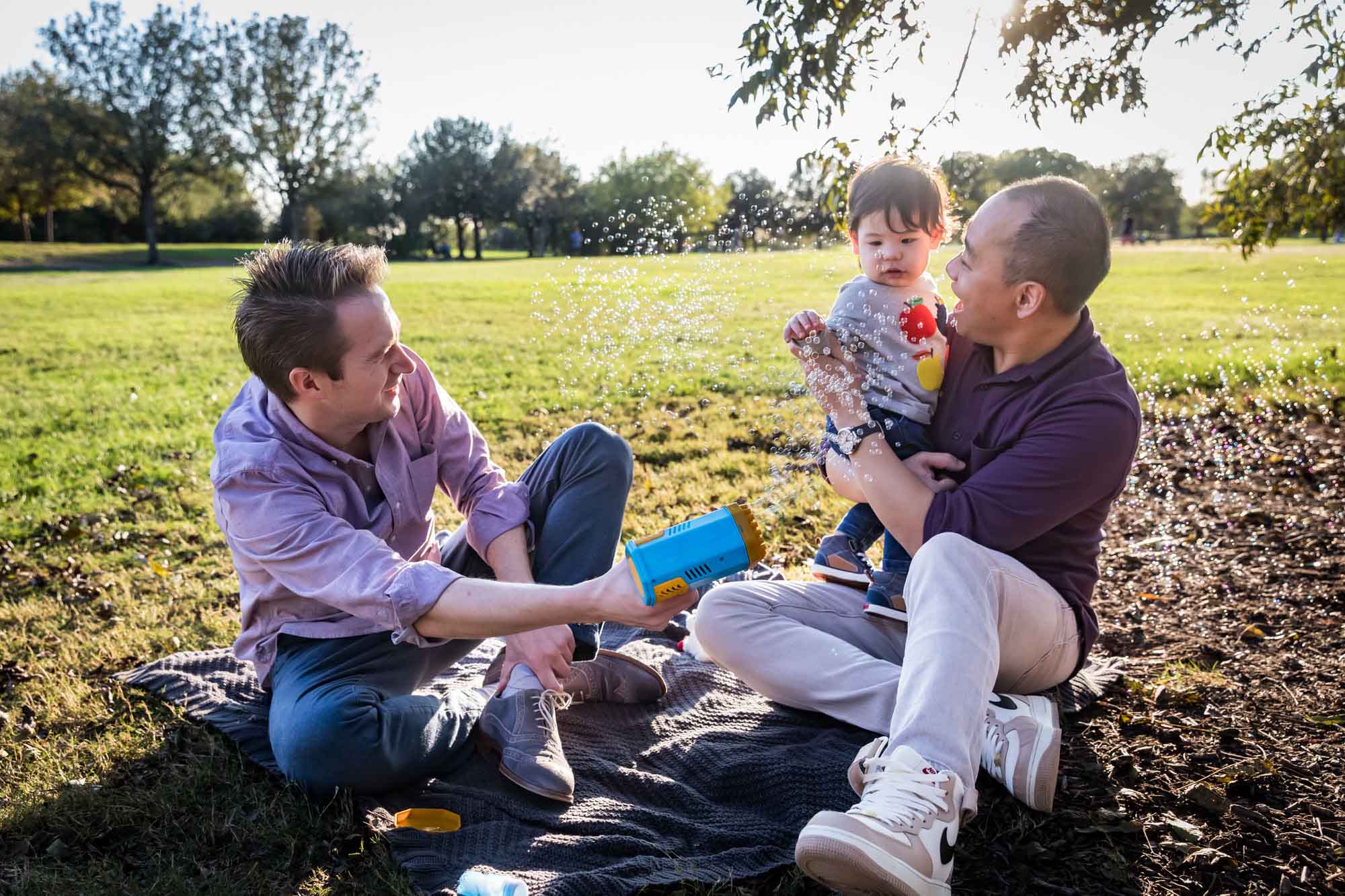Two fathers playing with little boy surrounded by bubbles while sitting on grass during a Mueller Lake Park family portrait