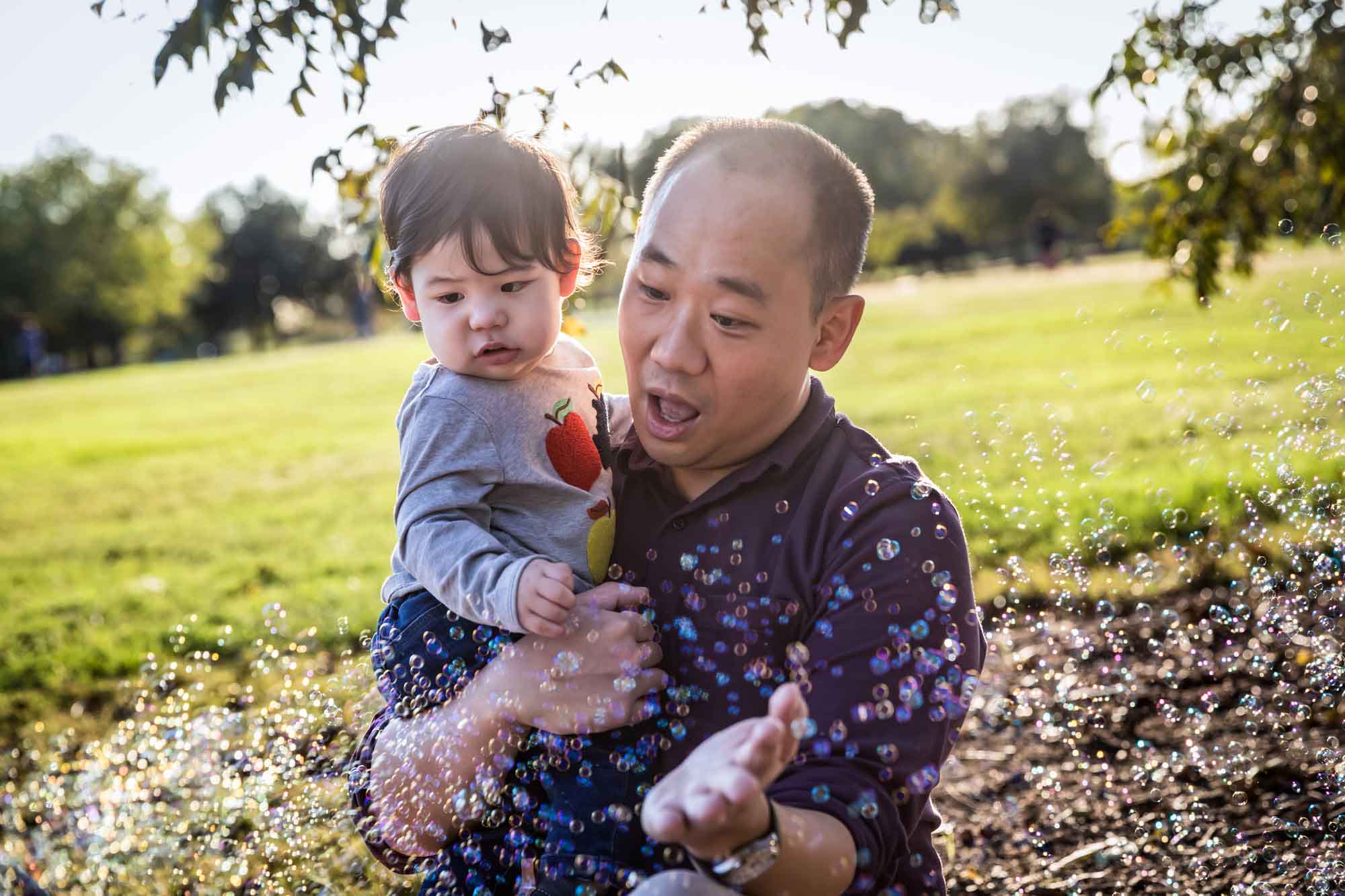 Father holding little boy surrounded by bubbles while sitting on grass during a Mueller Lake Park family portrait