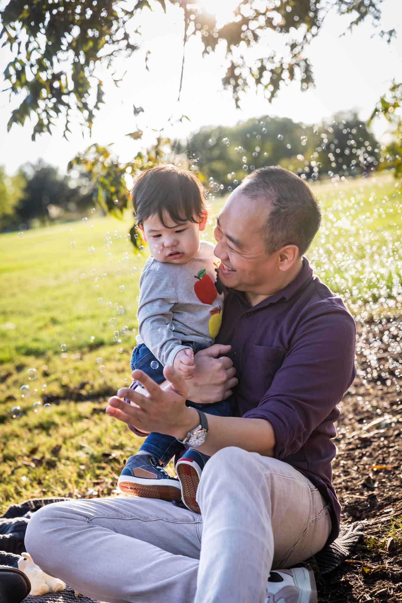 Father holding little boy surrounded by bubbles while sitting on grass during a Mueller Lake Park family portrait