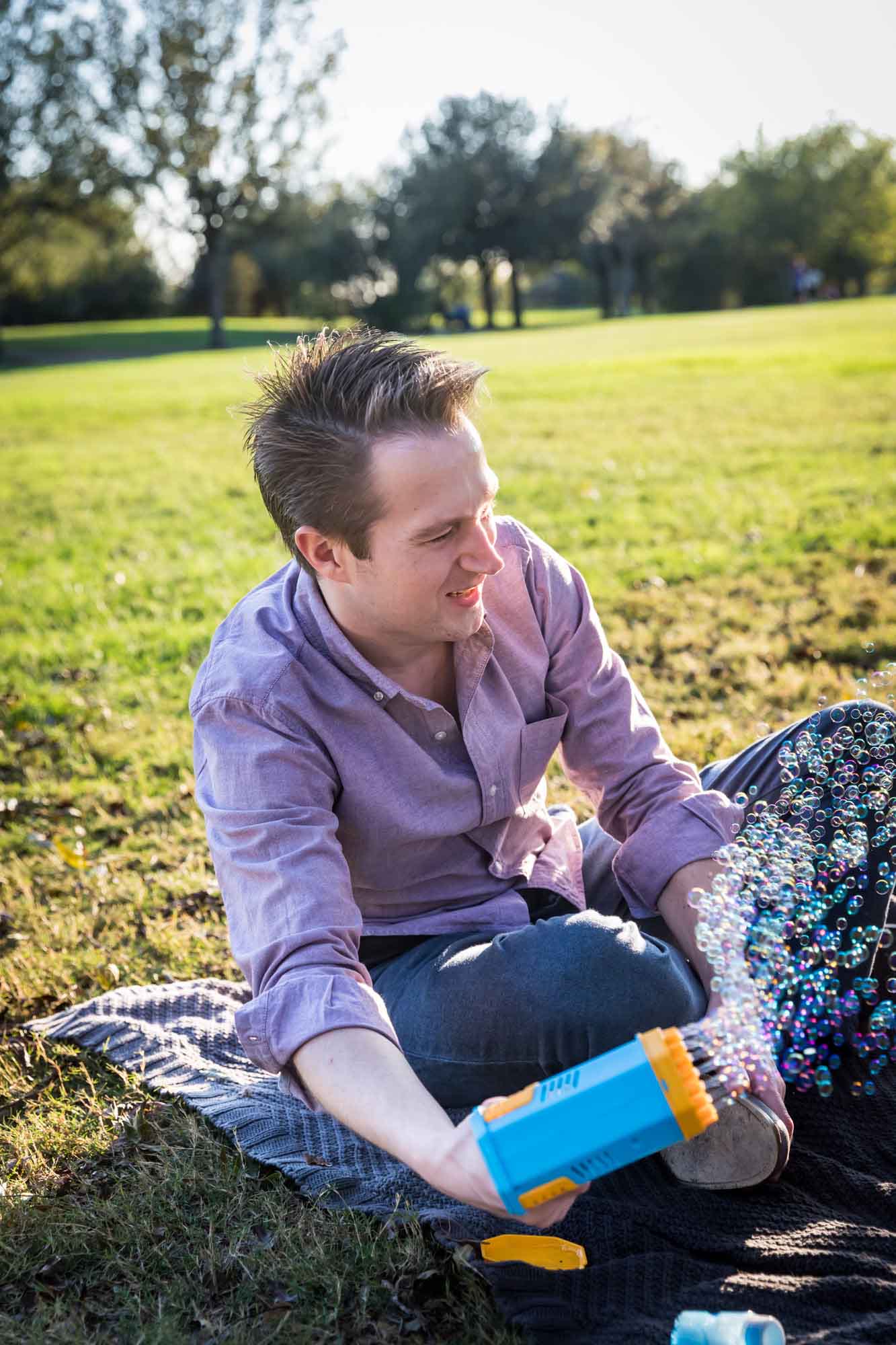 Father wearing pink shirt using machine to blow bubbles while sitting on grass during a Mueller Lake Park family portrait