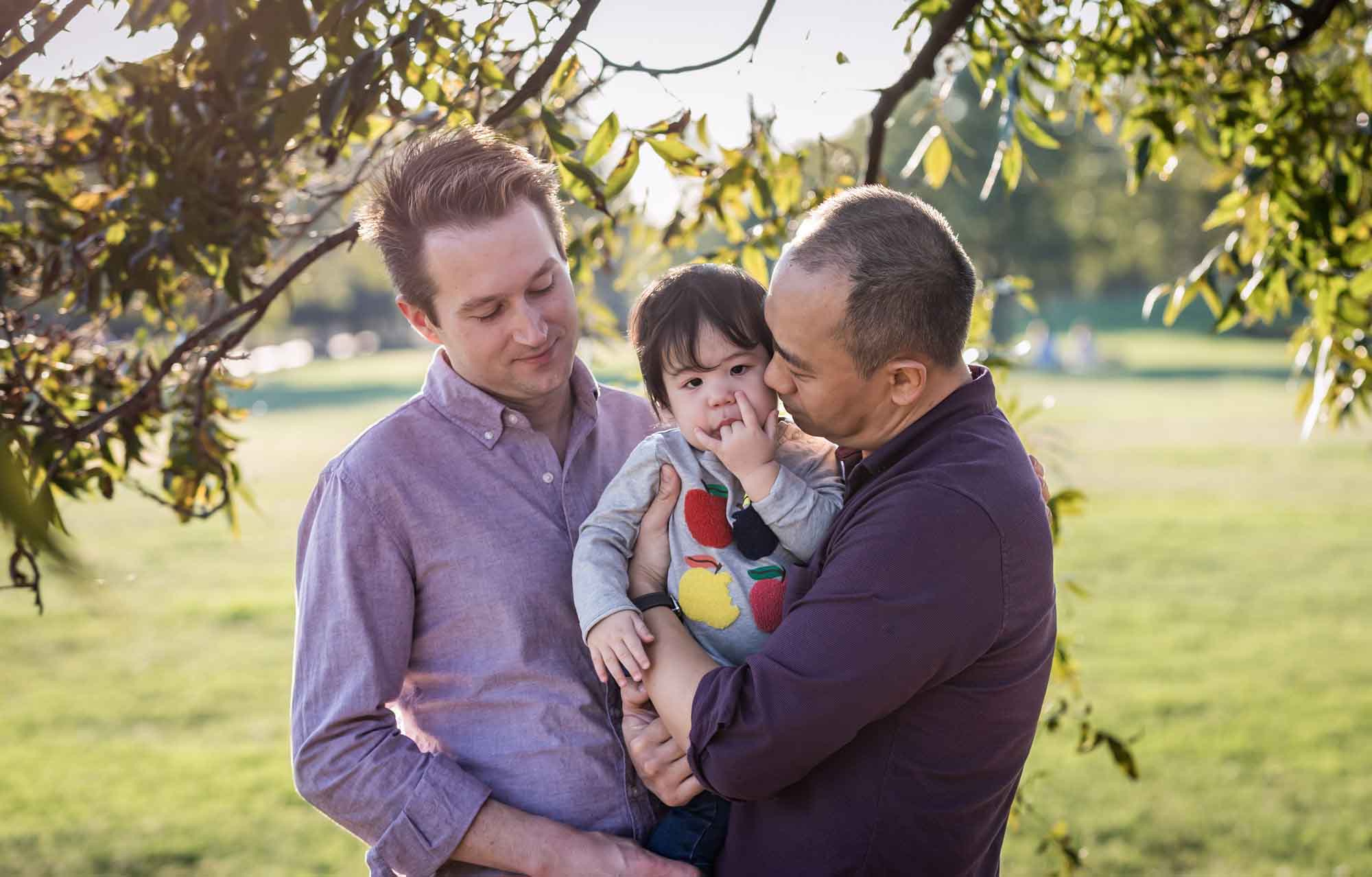 Two fathers holding little boy under trees in front of grass during a Mueller Lake Park family portrait