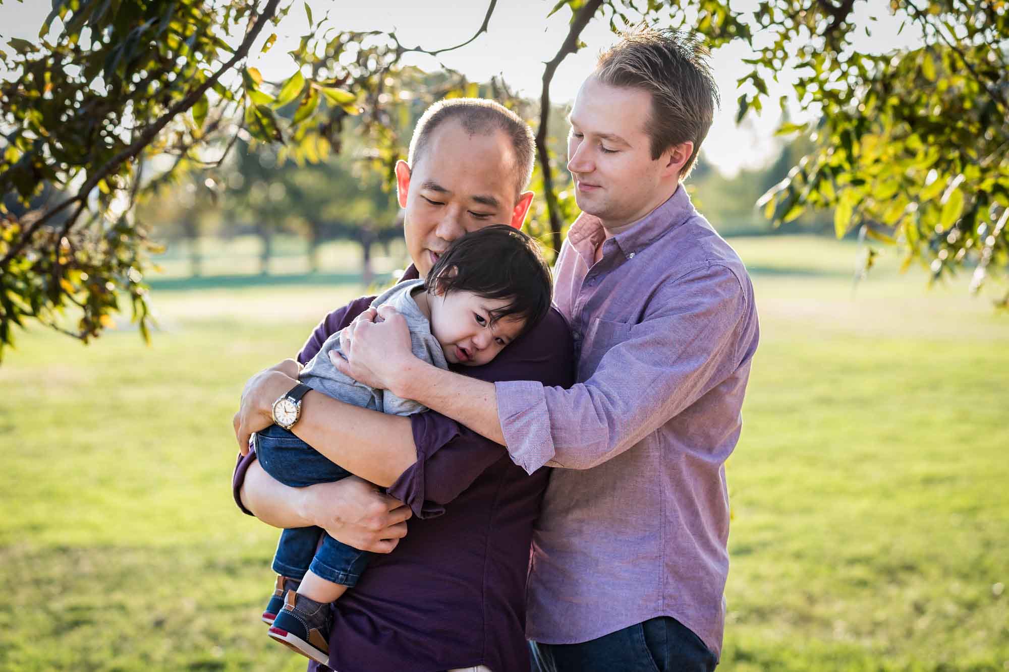 Two fathers holding little boy under trees in front of grass during a Mueller Lake Park family portrait