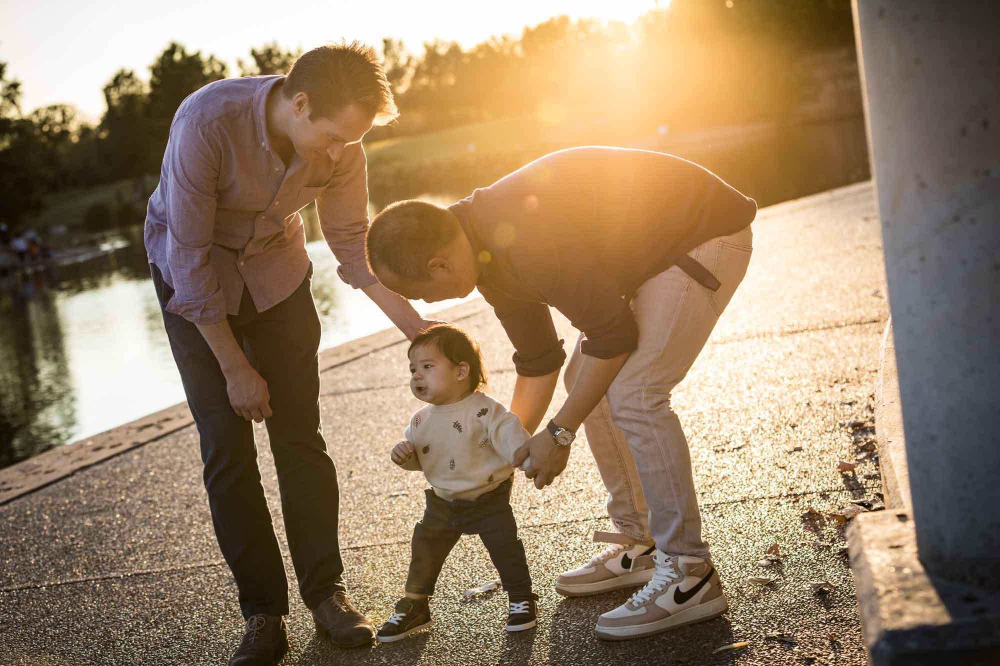 Little boy walking on ground with both hands held by two fathers during a Mueller Lake Park family portrait