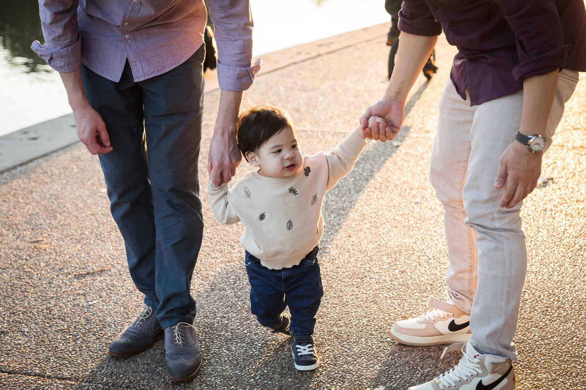 Little boy walking on ground with both hands held by two fathers during a Mueller Lake Park family portrait
