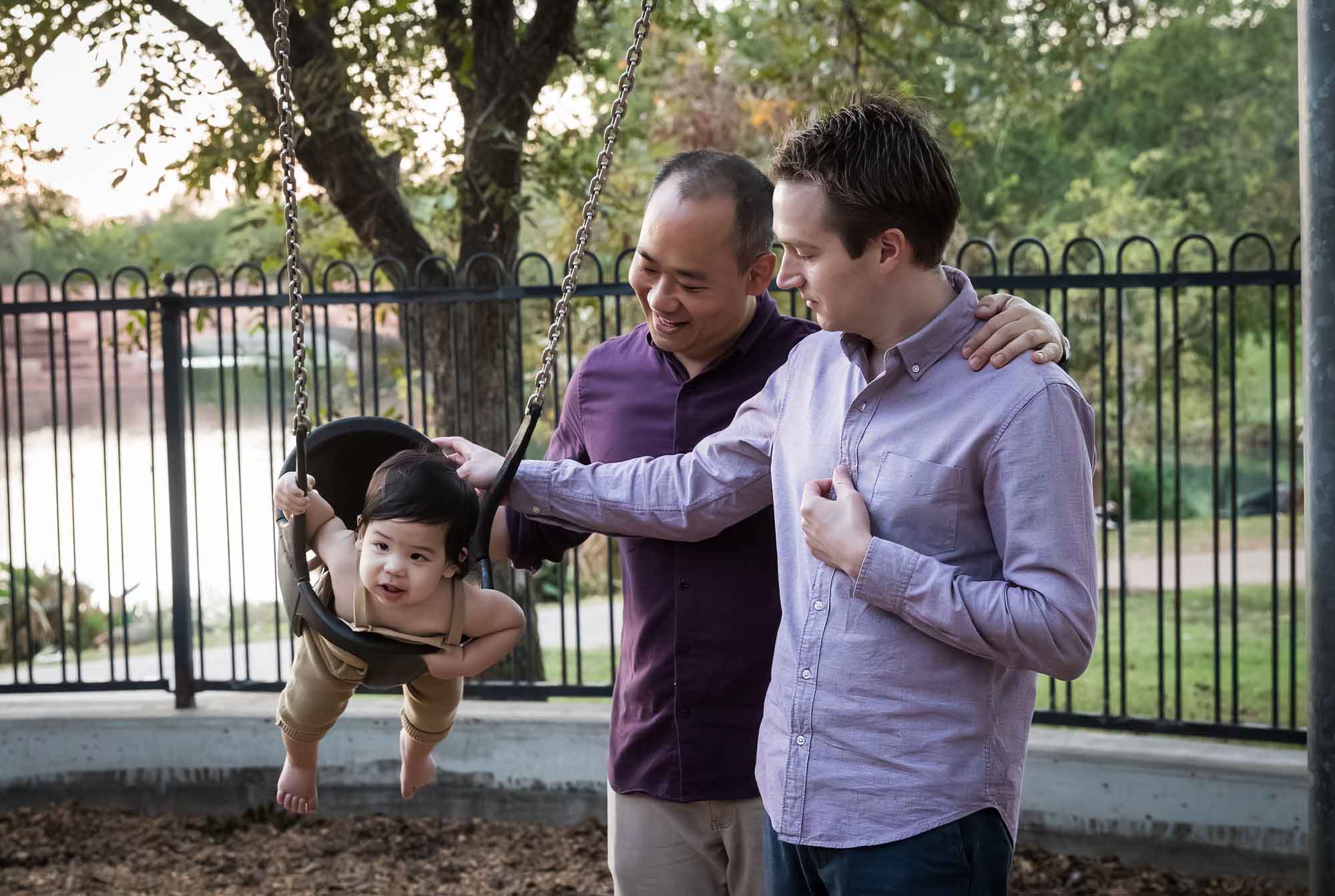 Two fathers pushing little boy on a swing at a playground during a Mueller Lake Park family portrait