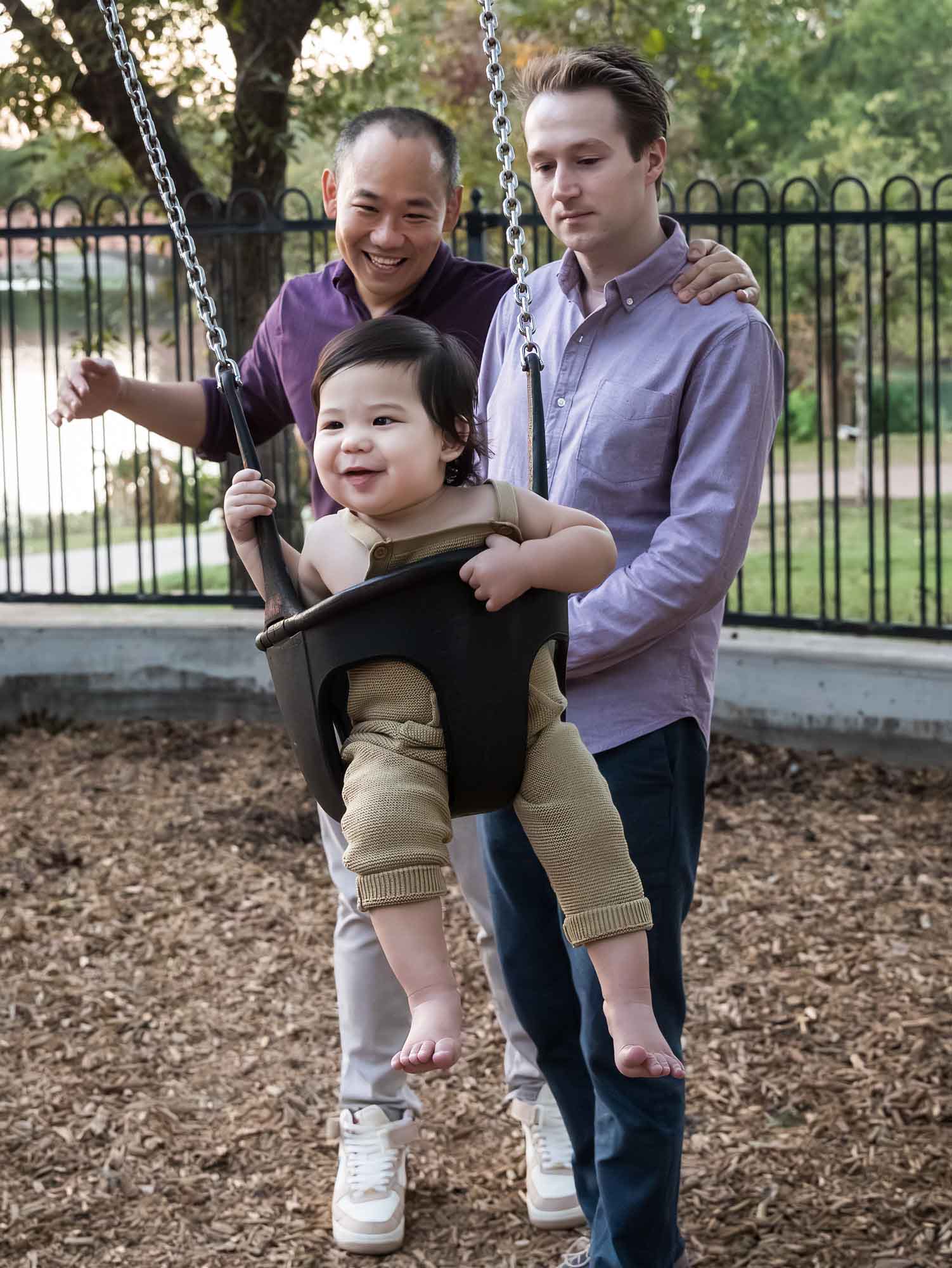 Two fathers pushing little boy on a swing at a playground during a Mueller Lake Park family portrait
