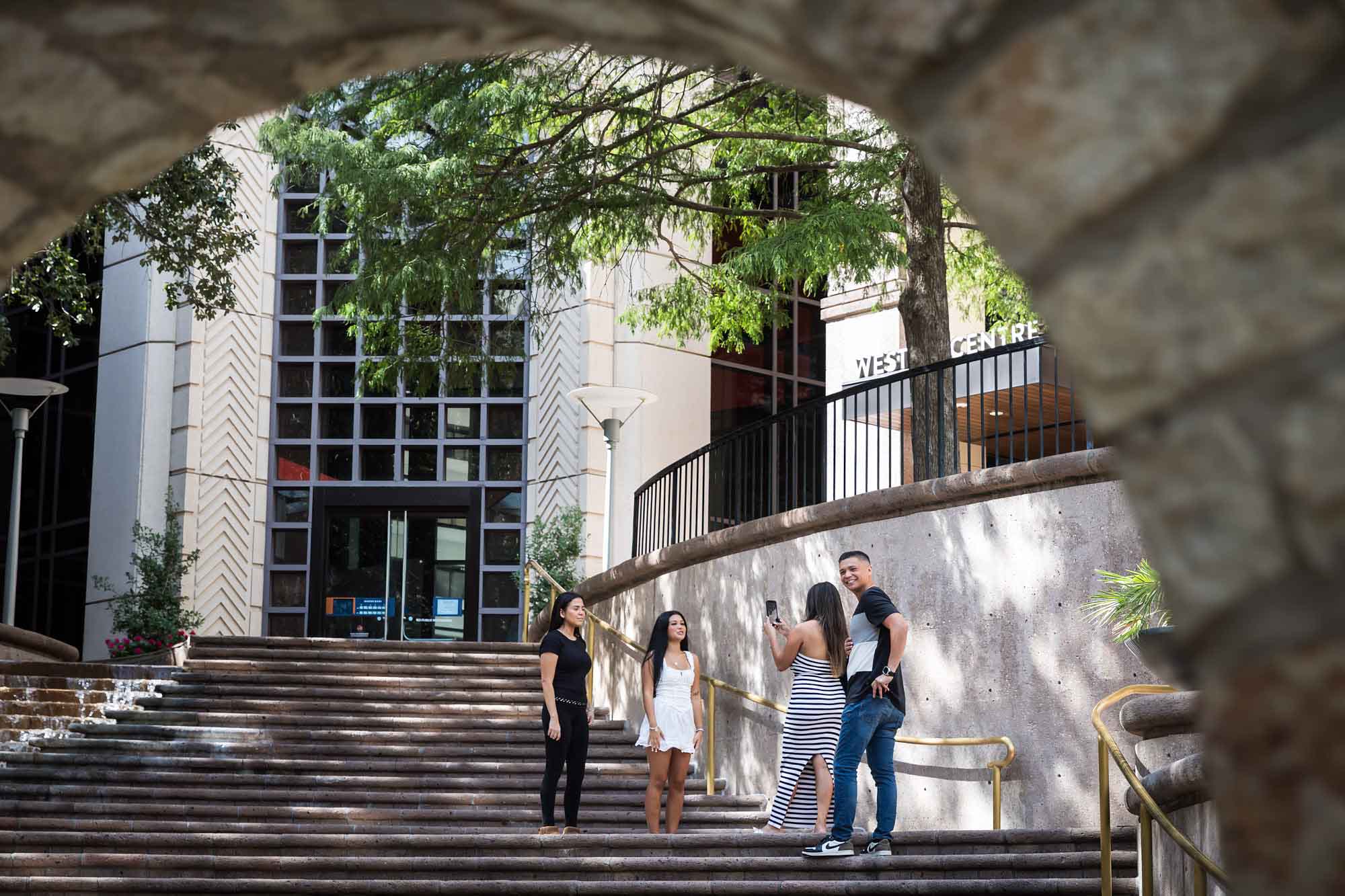 Two young girls and woman wearing striped dress holding phone in front of man wearing jeans on stairs during Riverwalk surprise proposal in San Antonio