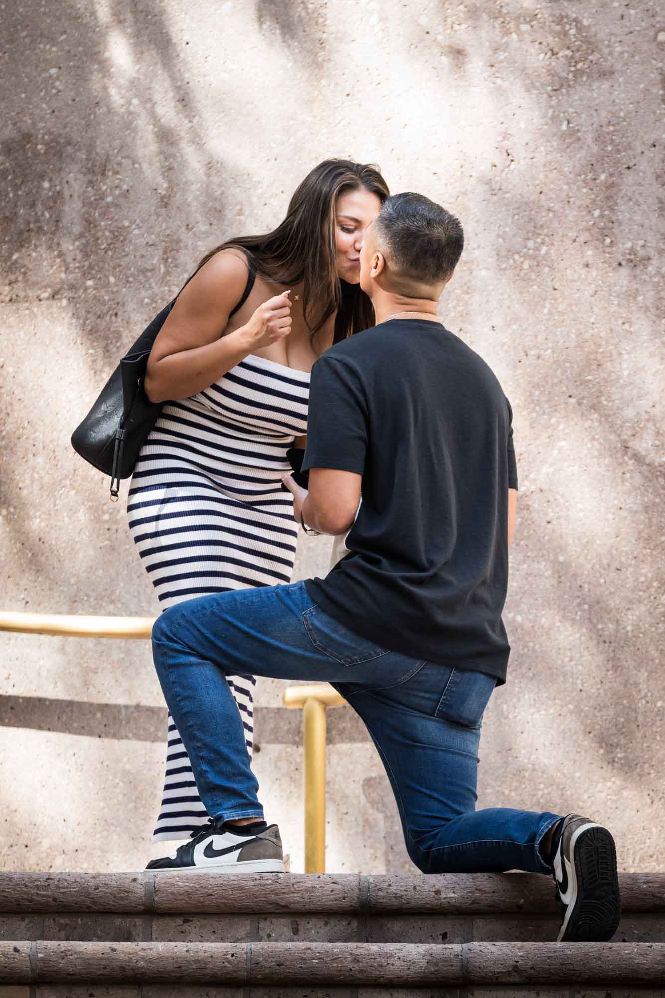 Woman wearing black and white striped dress bending down to kiss man bent down on knee on stairs during Riverwalk surprise proposal in San Antonio
