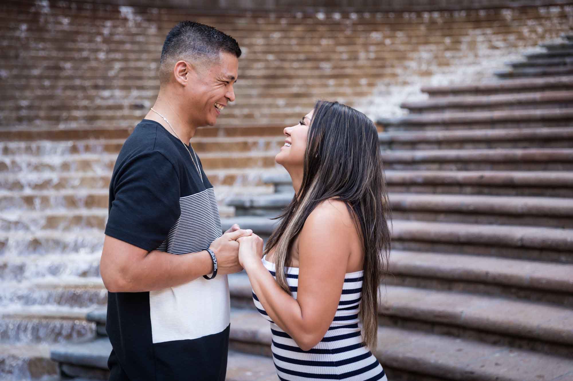 Woman wearing black and white striped, sleeveless dress with hand showing engagement ring holding hands with man wearing striped shirt and during a Riverwalk engagement photo shoot with waterfall and stairs in background