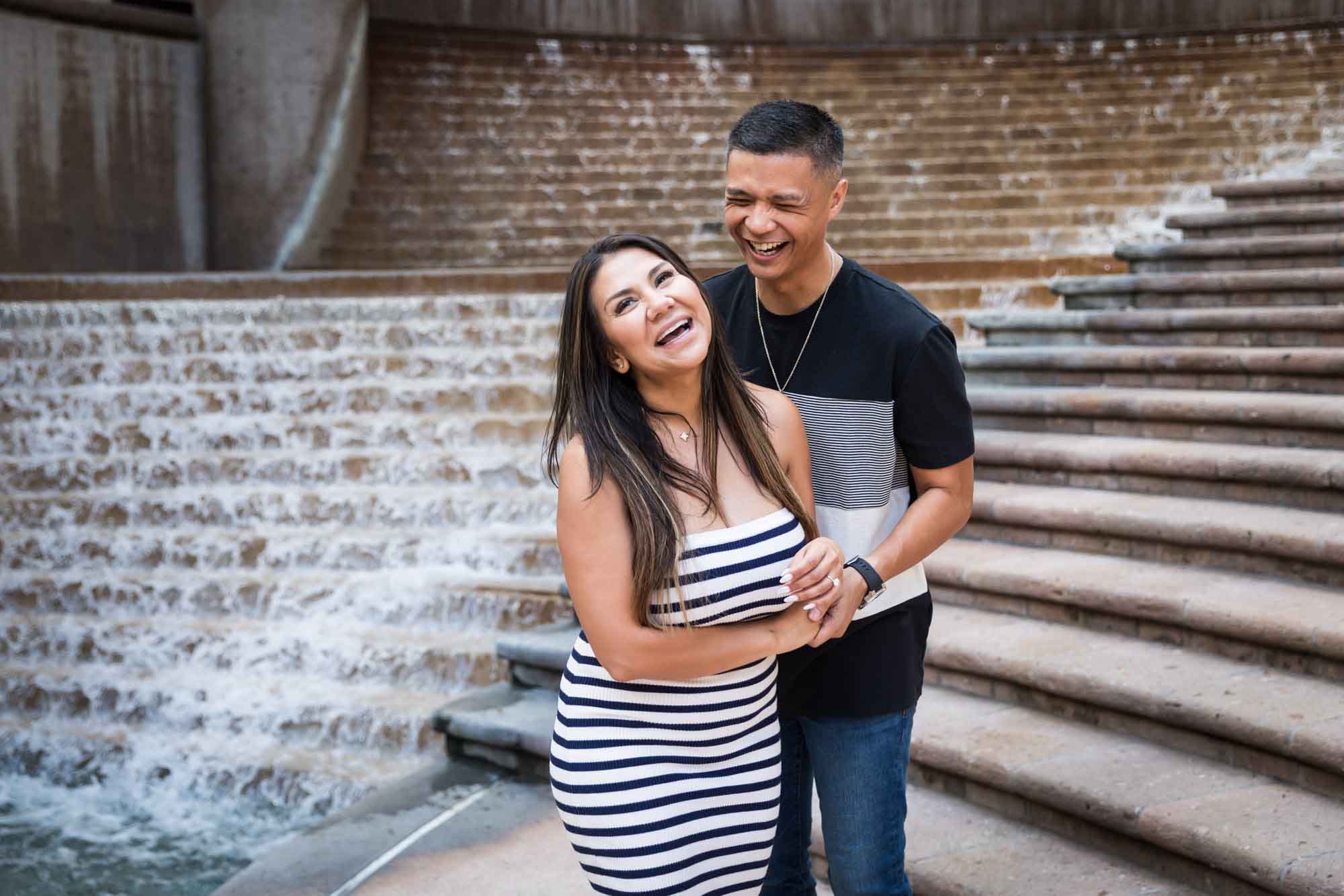 Woman wearing black and white striped, sleeveless dress with hand showing engagement ring standing in front of man wearing striped shirt and during a Riverwalk engagement photo shoot with waterfall and stairs in background