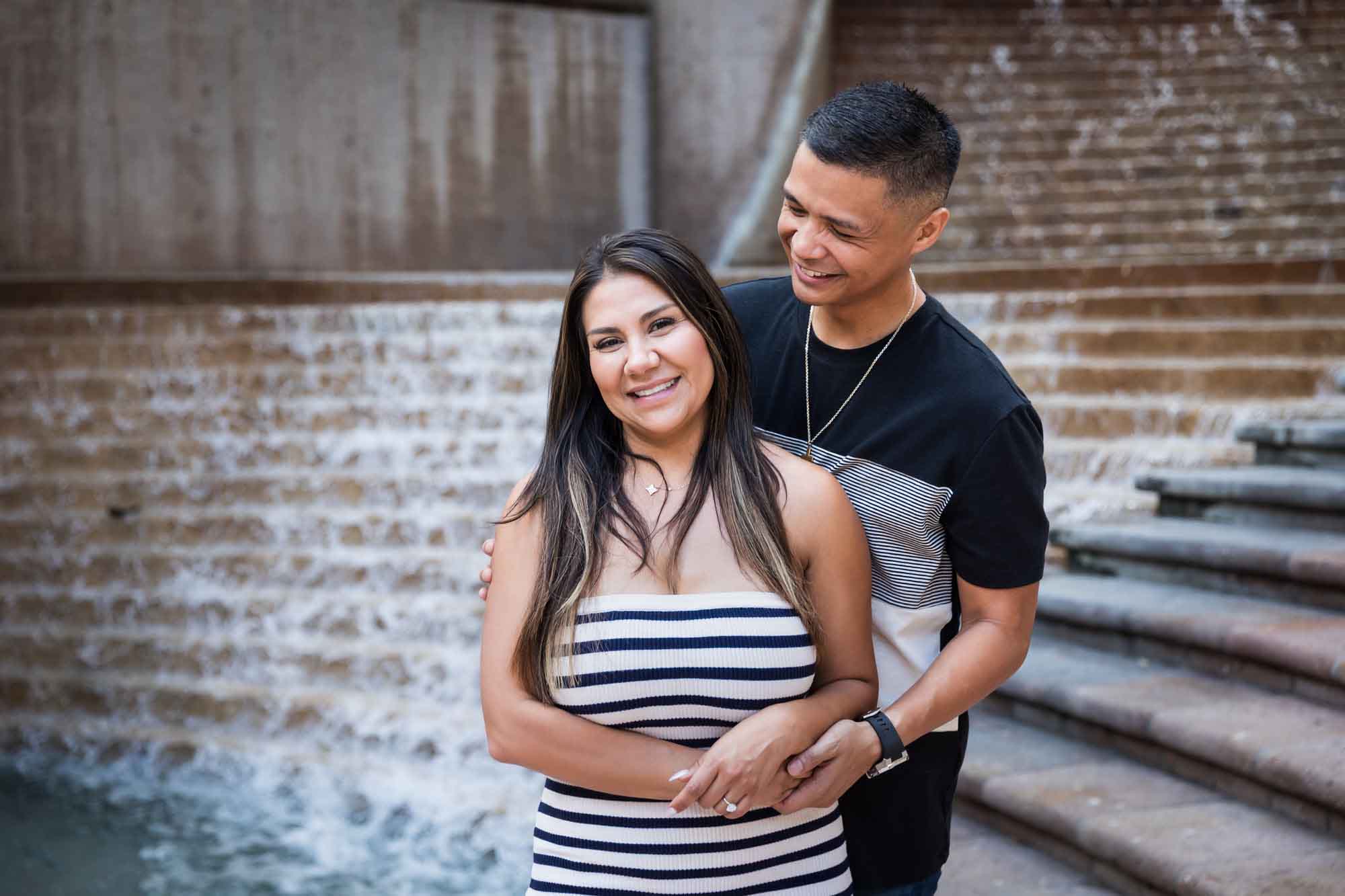 Woman wearing black and white striped, sleeveless dress with hand showing engagement ring standing in front of man wearing striped shirt and during a Riverwalk engagement photo shoot with waterfall and stairs in background