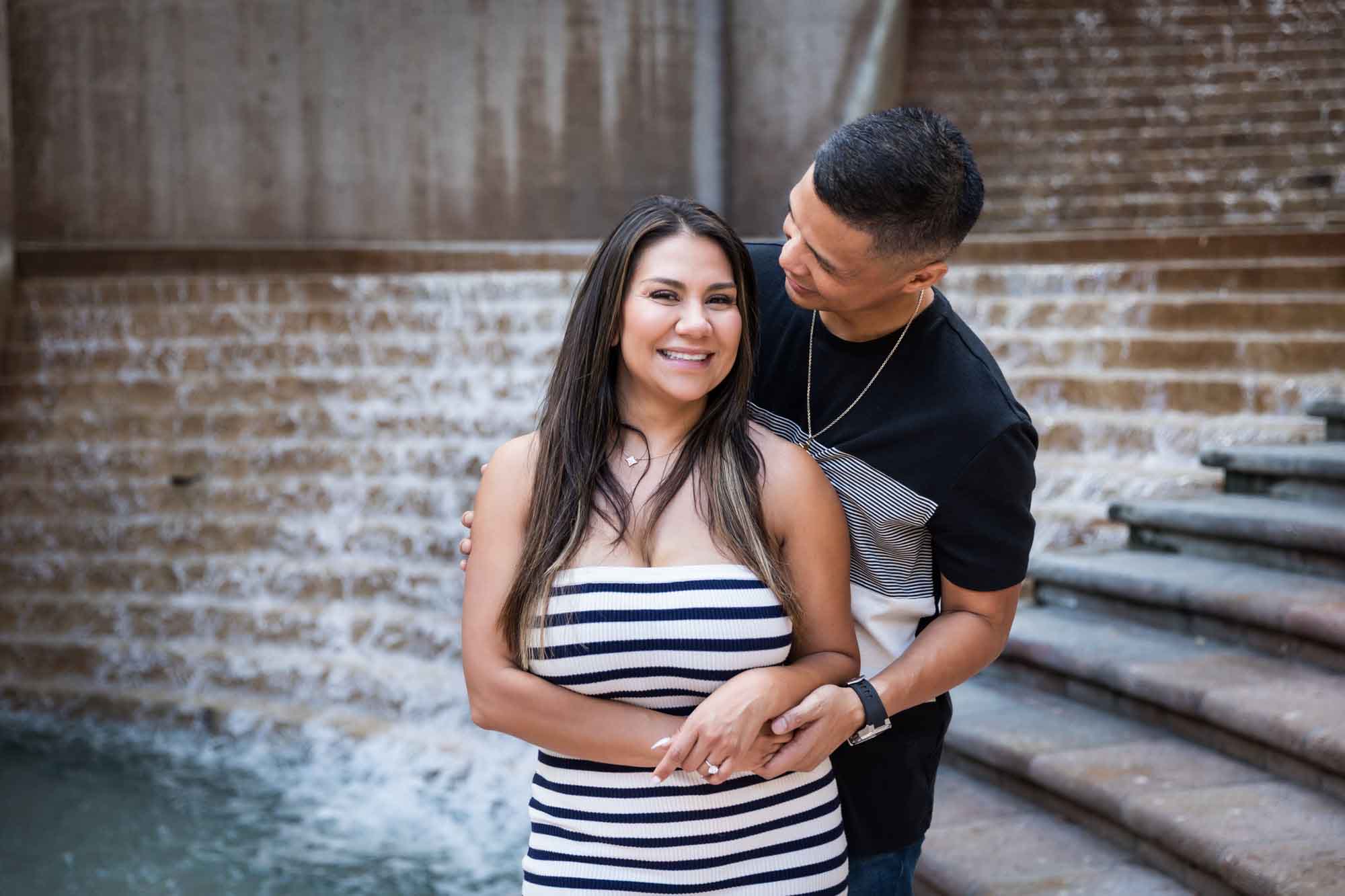 Woman wearing black and white striped, sleeveless dress with hand showing engagement ring standing in front of man wearing striped shirt and during a Riverwalk engagement photo shoot with waterfall and stairs in background