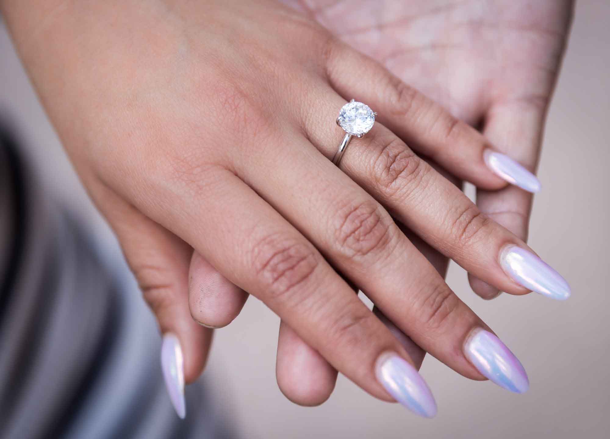 Close up of woman's hand showing round diamond engagement ring and purple iridescent nails resting on man's hands