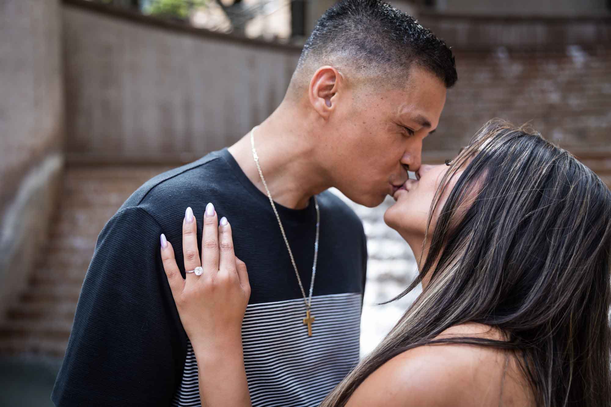 Woman wearing black and white striped, sleeveless dress with hand showing engagement ring resting on man wearing striped shirt kissing during a Riverwalk engagement photo shoot with waterfall and stairs in background